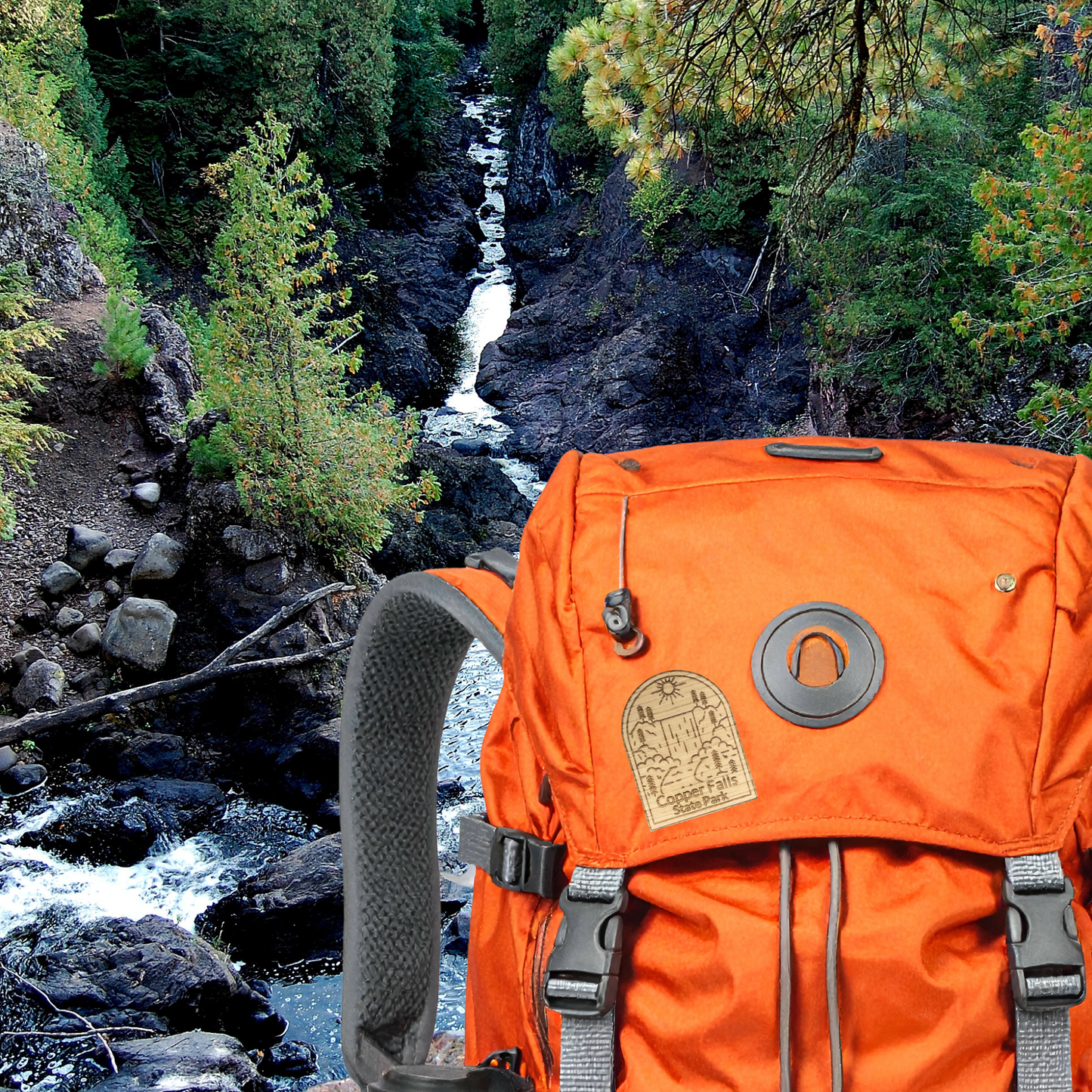 Orange backpack with a wood Copper Falls State Park pin attached in front of a scenic view of a river and trees
