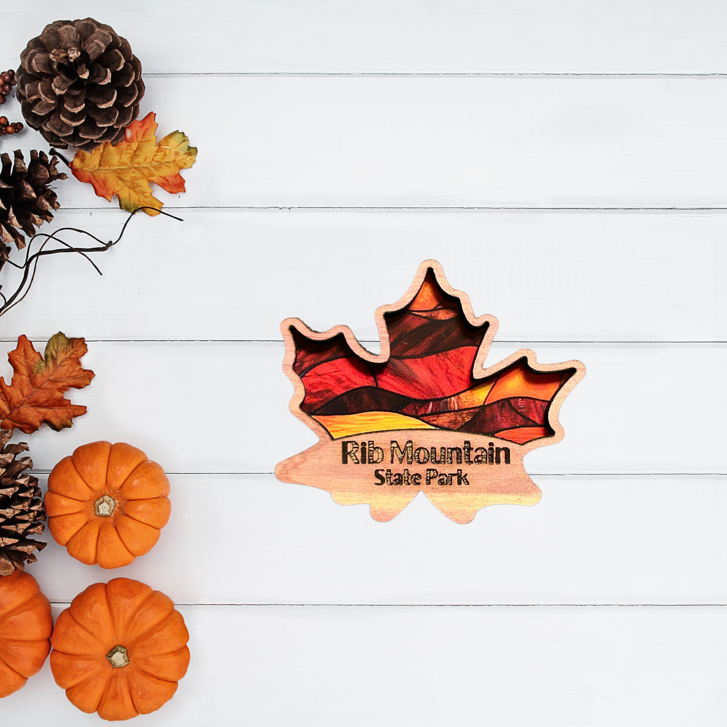 Cedar magnet in the shape of a maple leaf with a stained glass appearance and the Text “Rib Mountain State Park.” Photo background: pumpkins, pinecones and leaves.