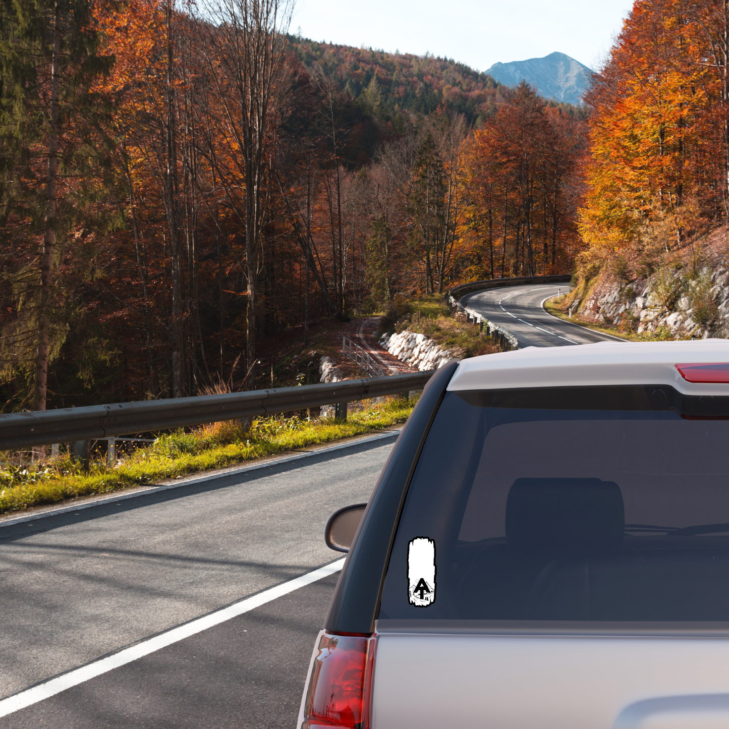 Weatherproof Appalachian Trail Blaze sticker with AT text and a scenic mountain design. Sticker attached to the back windshield of Car on a winding road with autumn trees and mountains in the background.