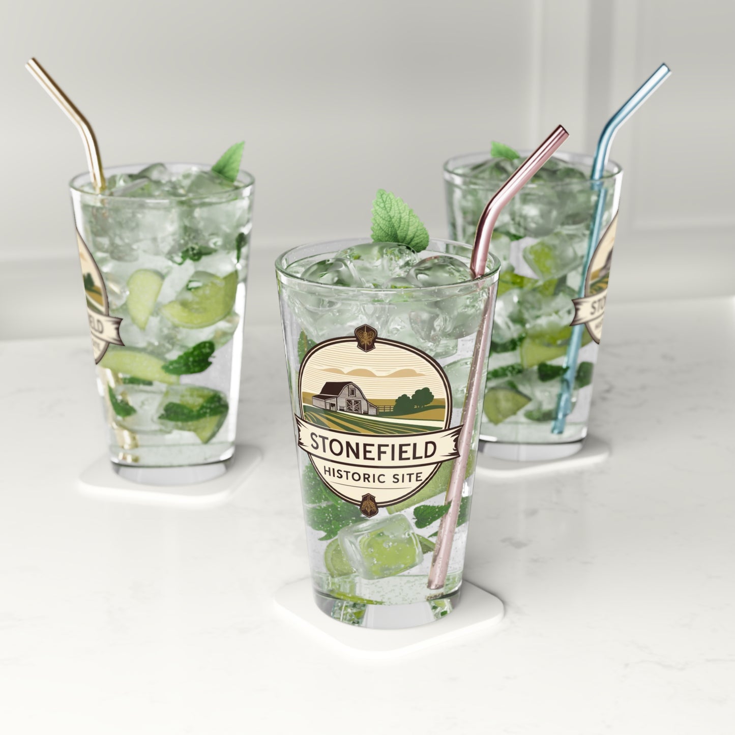 16oz pint glass featuring a logo design of a barn set amongst fields. Photo background: 3 glasses on a counter filled with water and lime wedges.