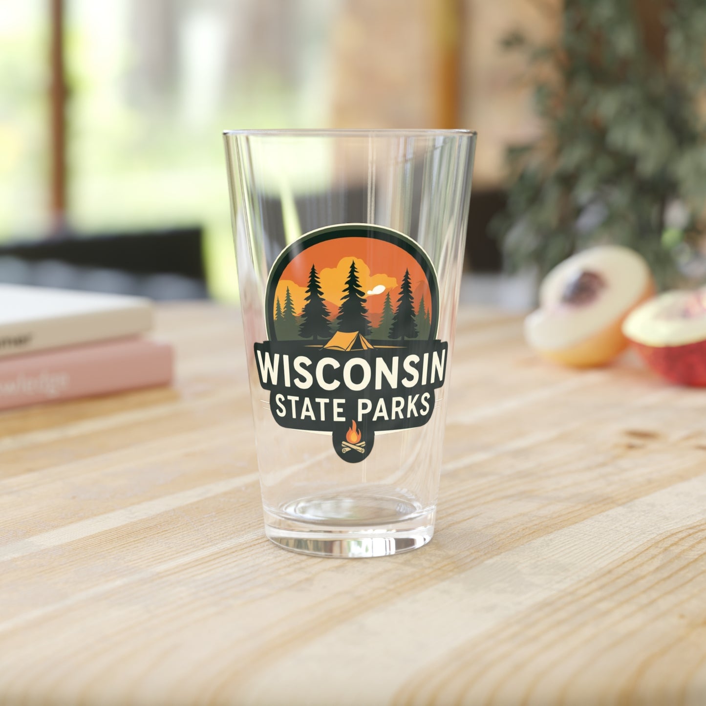 16 oz glass pint featuring a colorful design of a sunset over a campsite surrounded by pine trees and the text Wisconsin State parks. Photo background: glass sitting on a wood counter with books and fruit.