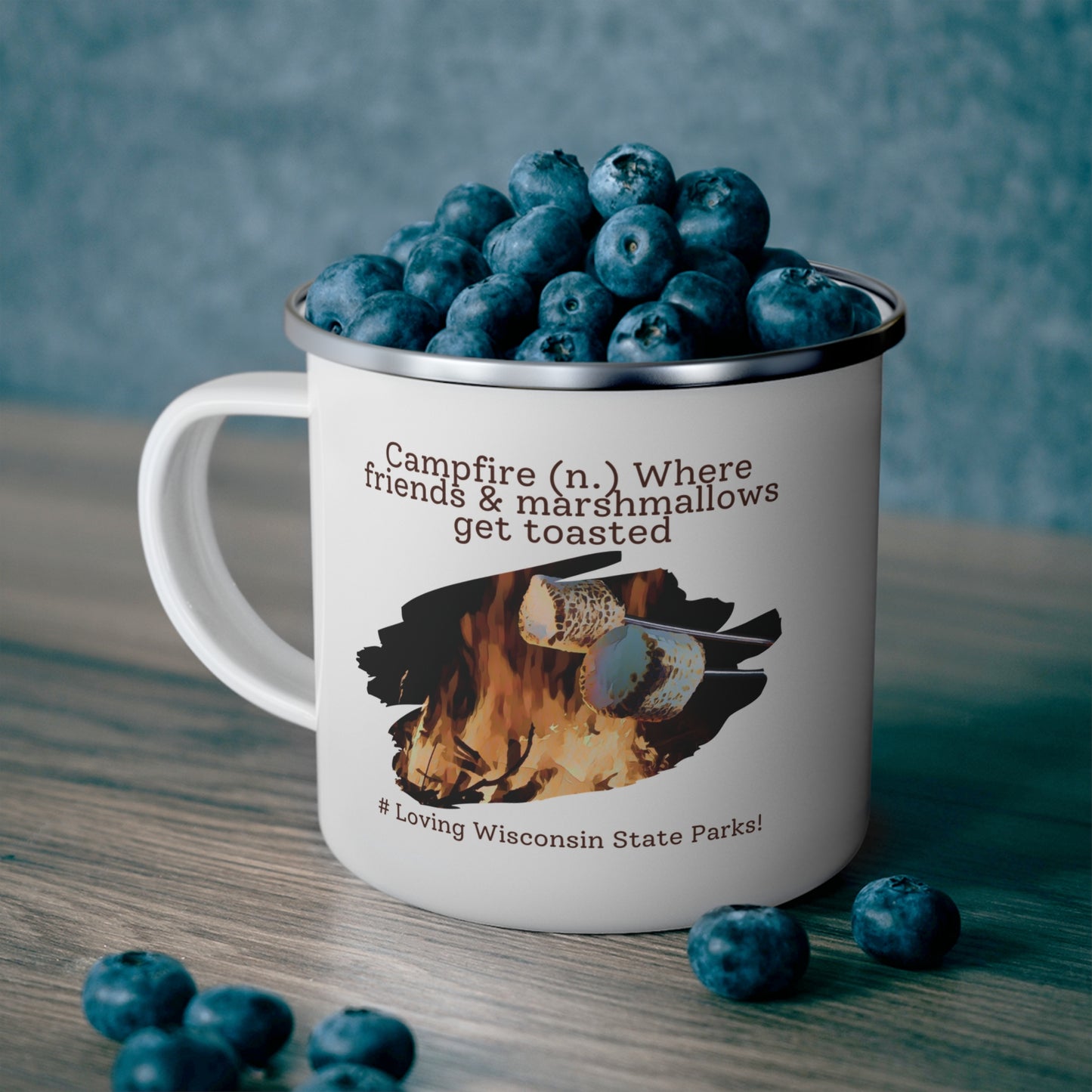 White enamel camper mug filled with blueberries sitting on a counter with a graphic design of a marshmallow being roasted over a fire and the text: campfire (n.) where friends and marshmallows get toasted. # loving Wisconsin State Parks”.