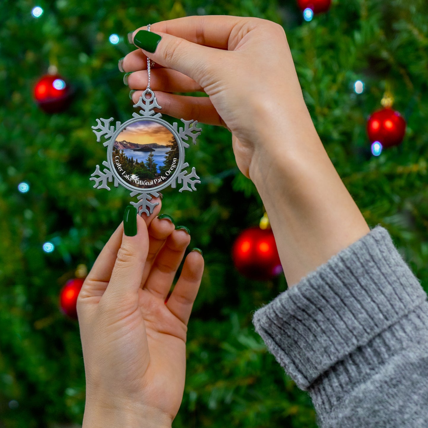 A person holding a pewter snowflake shaped ornament up against the background of a decorated Christmas tree. Ornament features a sunrise over Crater Lake design.