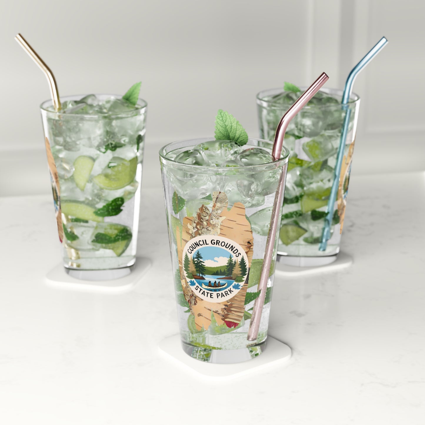 16 oz glass pint glass featuring a birch bark design with a depiction Native Americans in a birch bark canoe paddling on a river. Photo Background: three glasses sitting on a counter filled with water and lime slices.