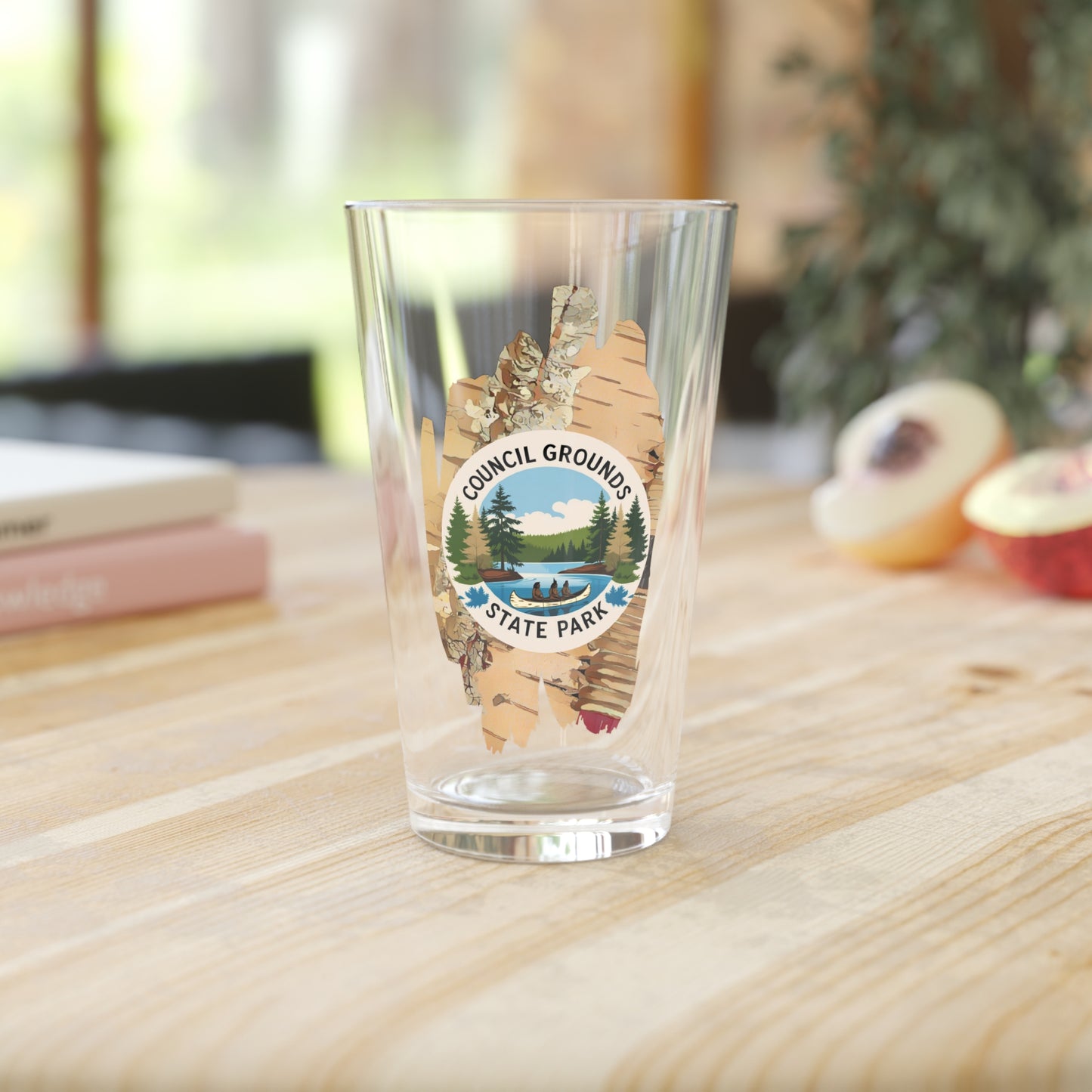 16 oz glass pint glass featuring a birch bark design with a depiction Native Americans in a birch bark canoe paddling on a river. Photo Background: glass sitting on a wood counter with books and fruit.