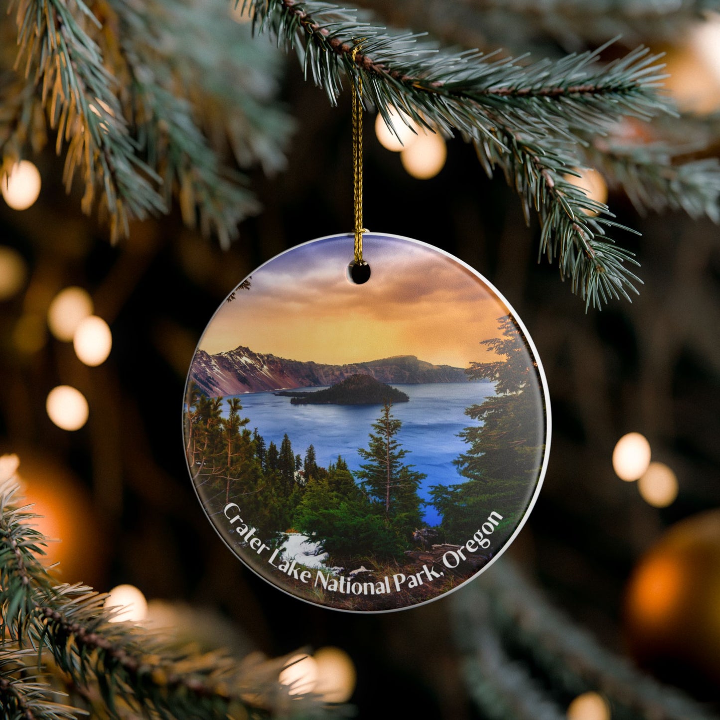 Round ceramic ornament featuring a vivid sunrise over Crater Lake design. Photo background: ornament hanging from a Christmas tree branch.