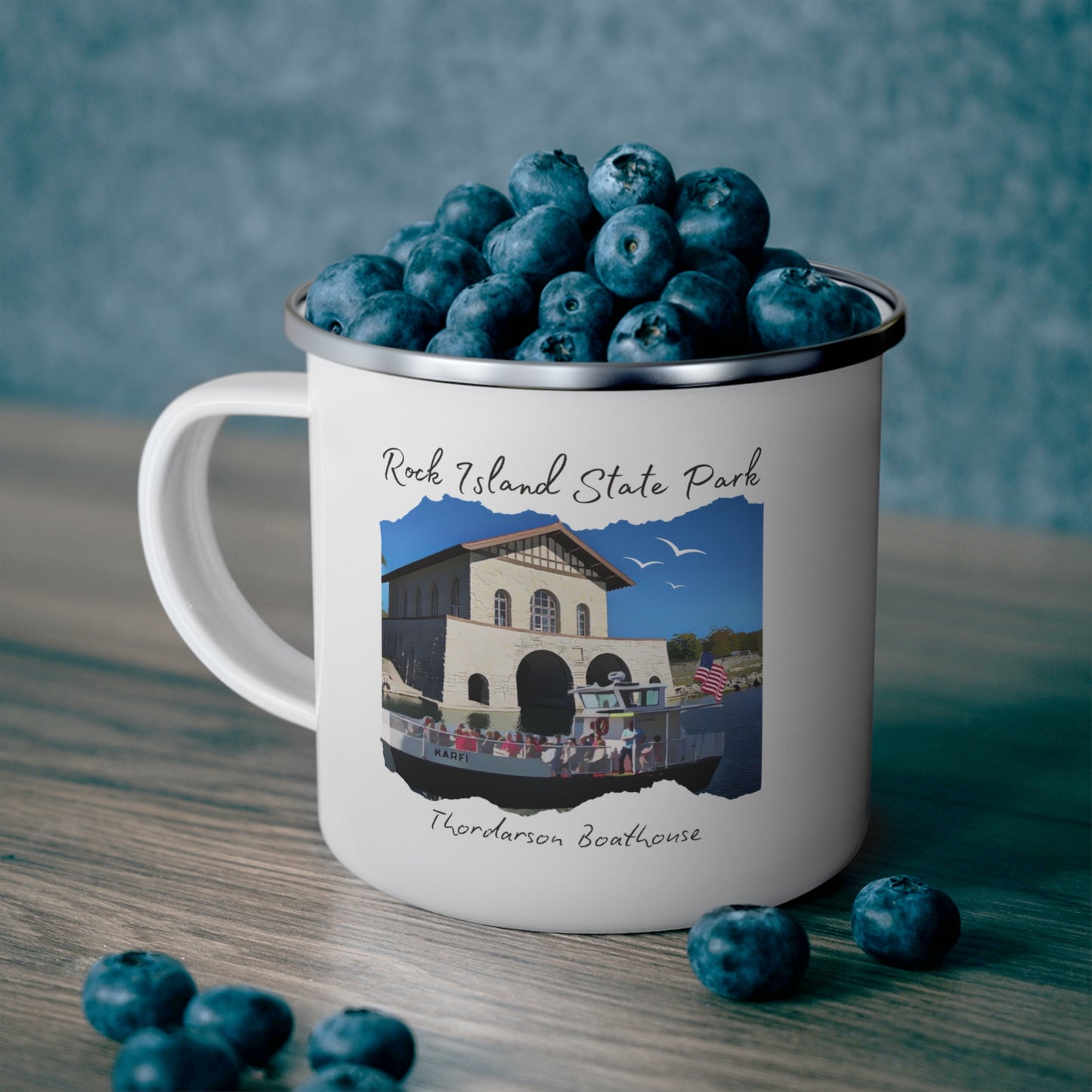 White enamel campfire mug depicting a scene of a ferry boat pulling up to the stone boat house on Rock Island. Photo background: mug full of blueberries sitting on a counter.