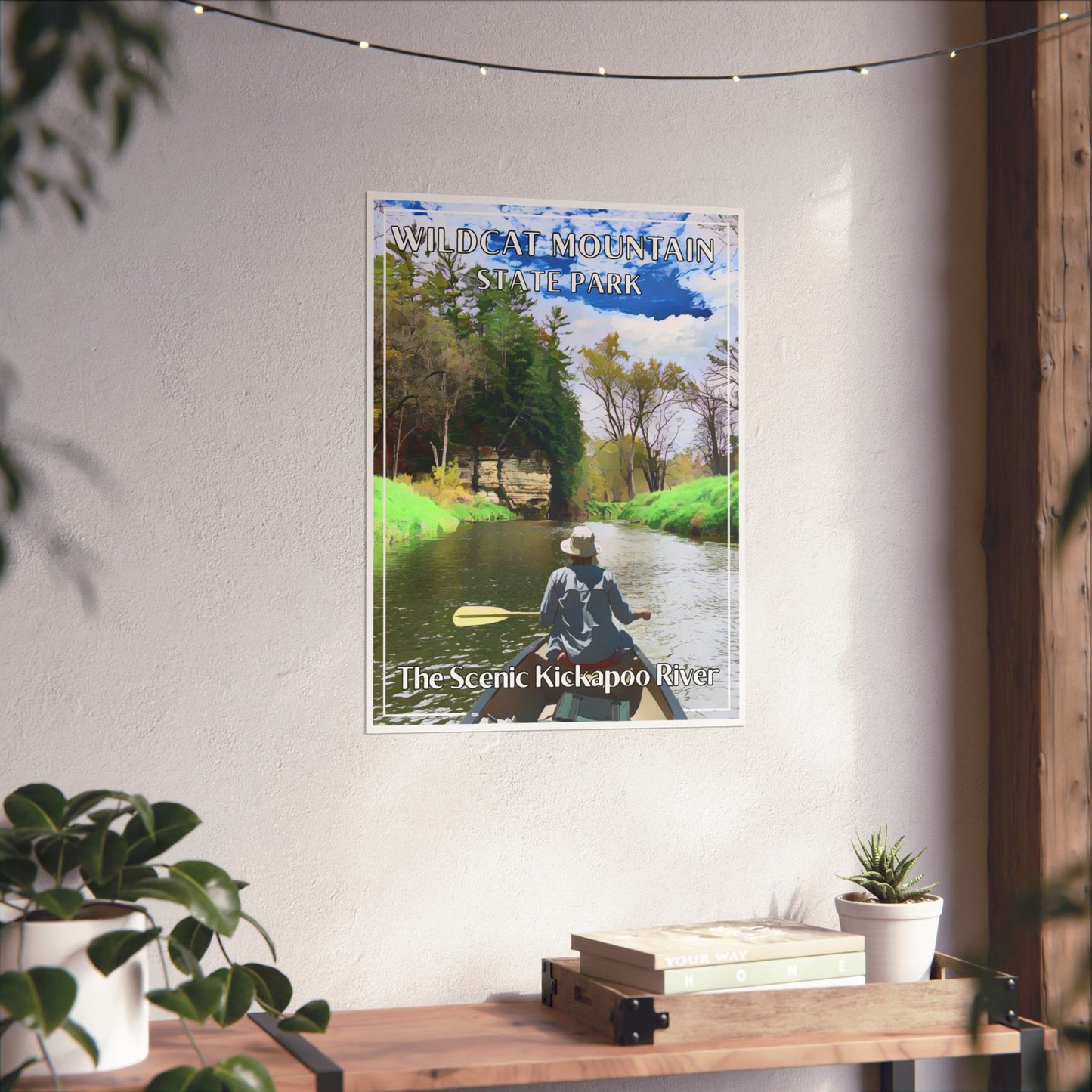 Poster featuring a canoeist paddling down a scenic river lined with trees and rock outcroppings. Photo background: poster hanging on a wall above a wood shelf with books and plants.