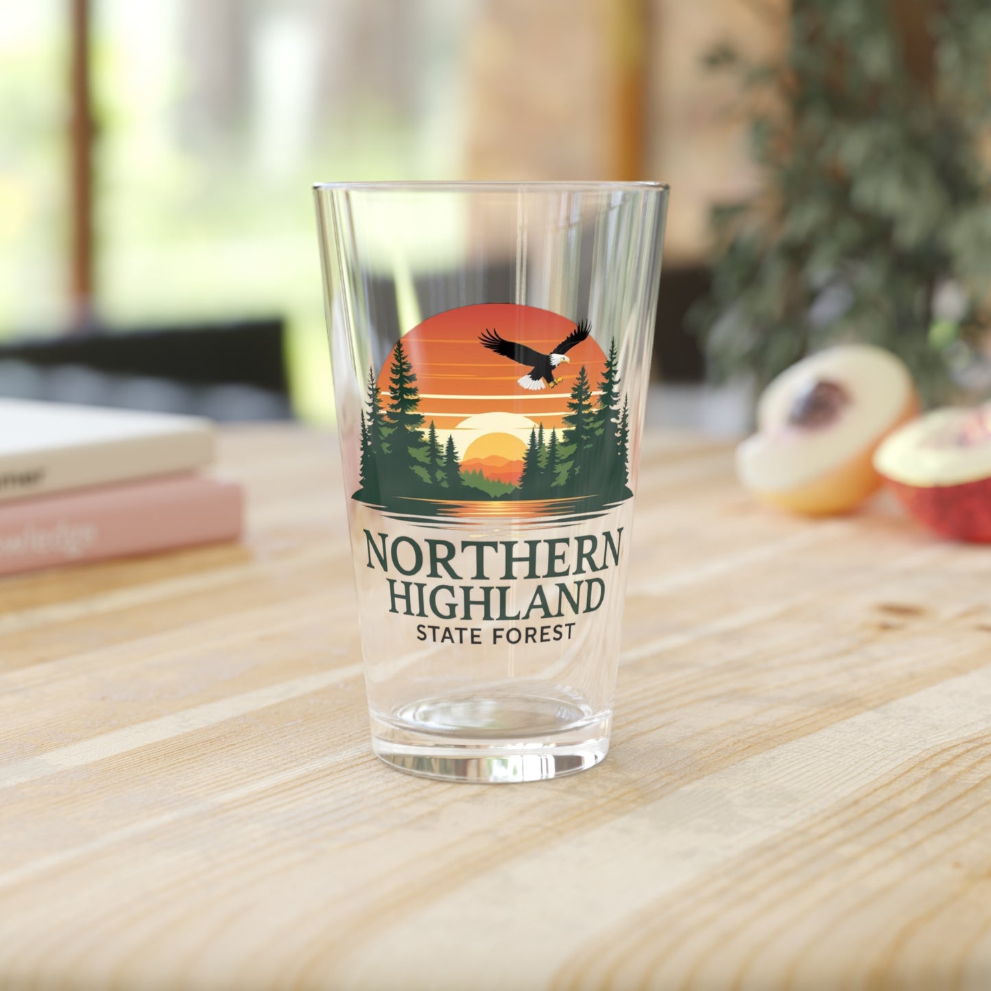 16 oz glass pint featuring a colorful design of a sunset over a lake surrounded by pine trees and a Bald Eagle Soaring overhead, with the text Northern Highland State Forest. Photo background: glass sitting on wood counter with books and fruit.