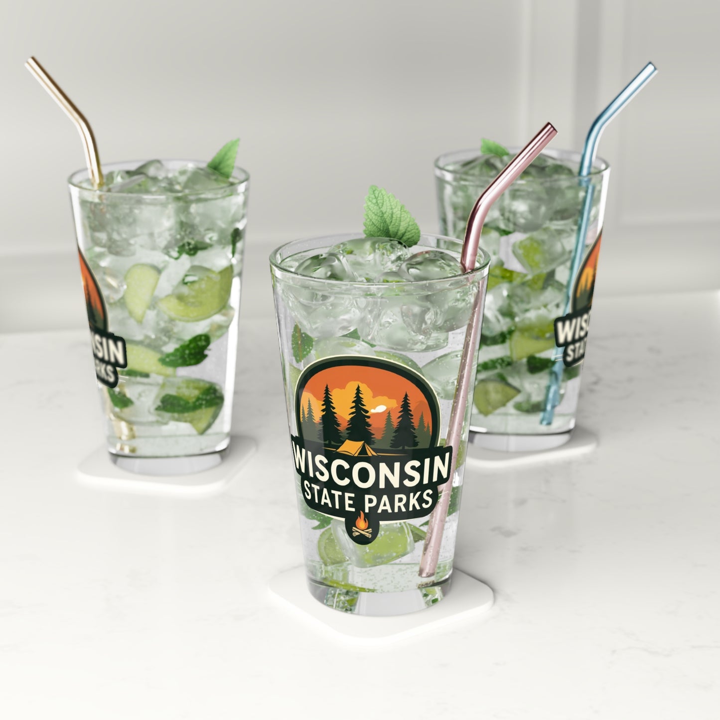 16 oz glass pint featuring a colorful design of a sunset over a campsite surrounded by pine trees and the text Wisconsin State parks. Photo background: three glasses sitting on a counter filled with water and lime wedges.