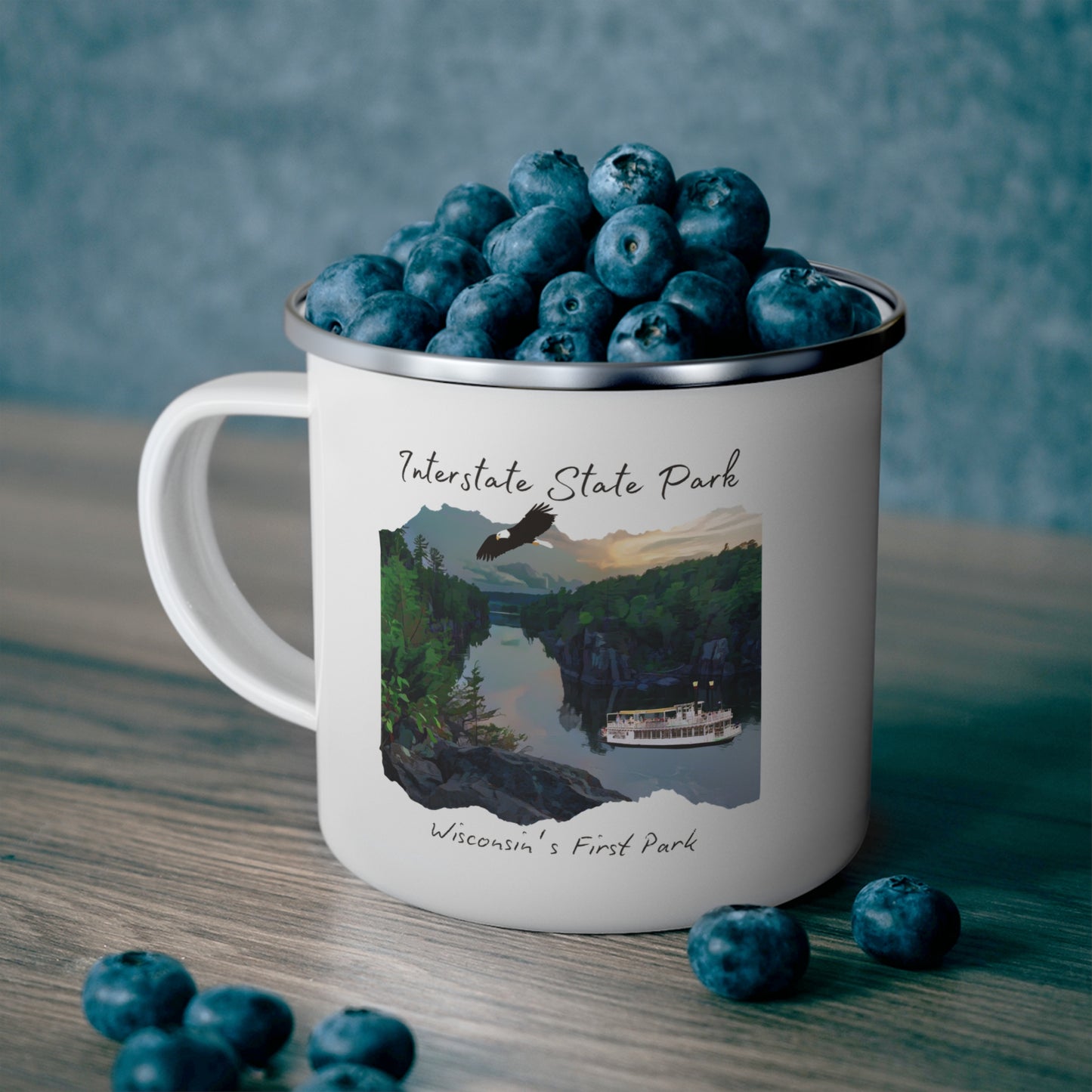 White enamel mug depicting the St Croix River at sunset with an eagle flying above and a paddle wheel boat on the river. Photo background: mug sitting on a shelf full of blueberries.