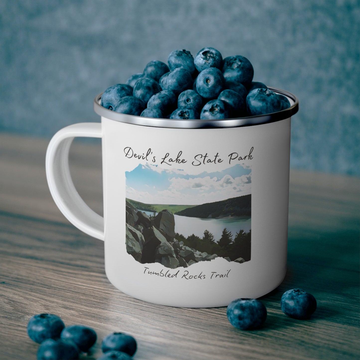 White enamel campfire mug with picture overlooking Devil’s Lake and the text Devil’s Lake State Park, Tumbled Rocks Trail. Background: mug sitting on counter with blueberries.