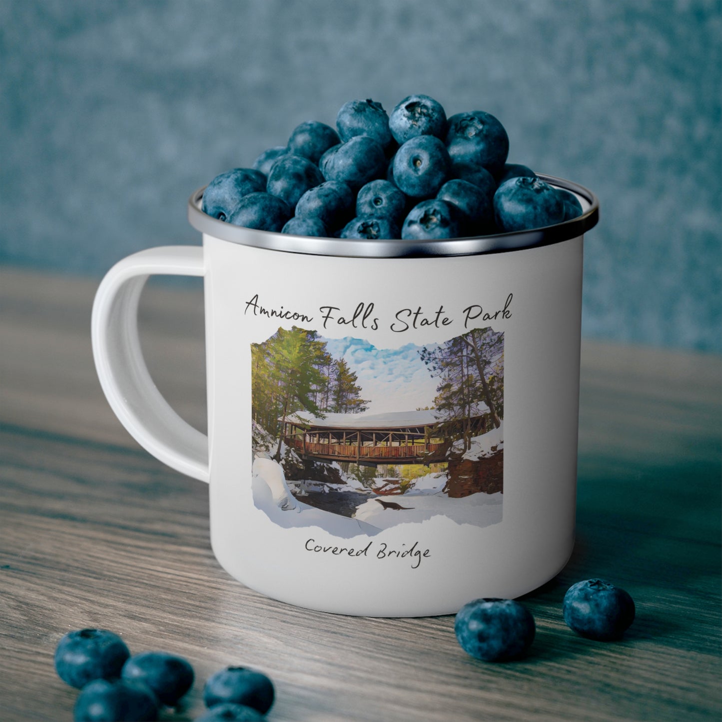 White enamel mug depicting the covered bridge at Amnicon Falls State Park in winter. Background: mug sitting on a counter, full of blueberries.