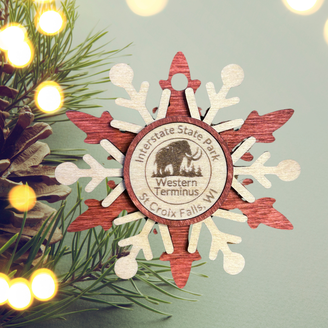 Red snowflake wood ornament depicting a woolly mammoth. Photo background: evergreen branch with lights and a pinecone.