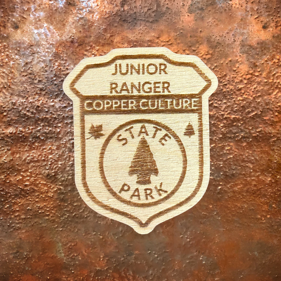 Wood Shaped badge with the text “Junior Ranger Copper Culture State Park” and an arrowhead symbol. Photo background: copper metal.
