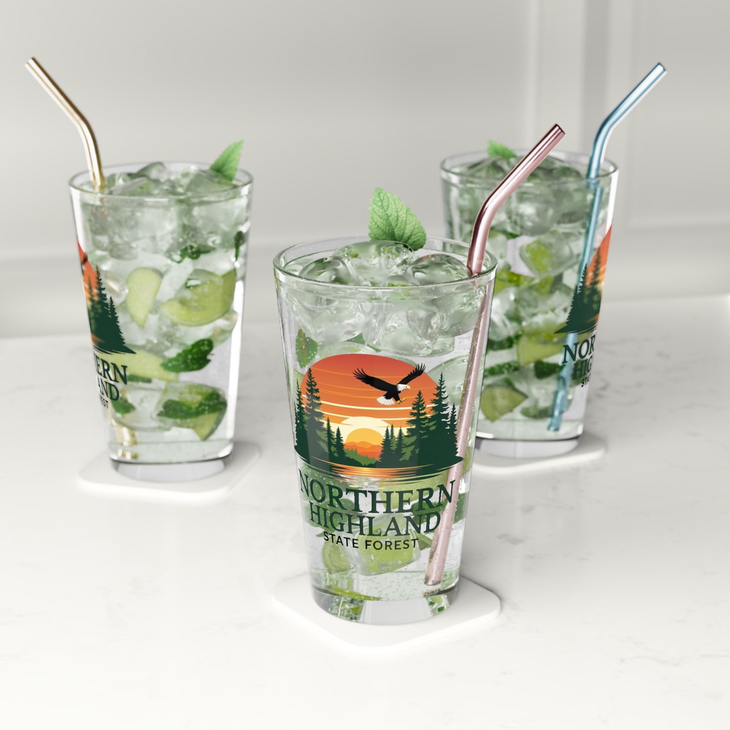 16 oz glass pint featuring a colorful design of a sunset over a lake surrounded by pine trees and a Bald Eagle Soaring overhead, with the text Northern Highland State Forest. Photo background: three glasses sitting on the counter filled with water and lime wedges.