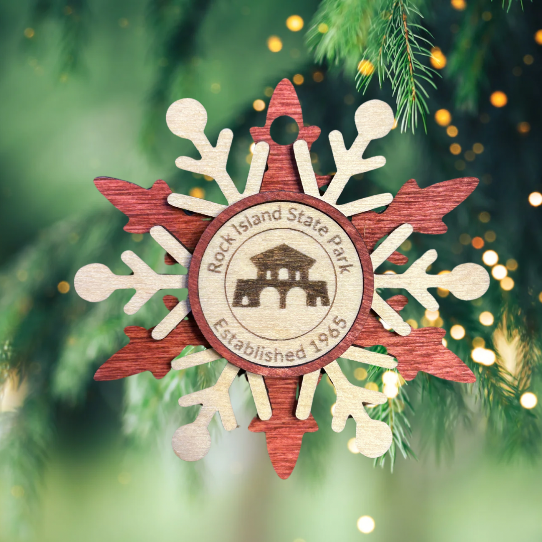 Red snowflake wood ornament depicting the boat house at Rock Island State Park. Photo background: evergreen branch with lights.
