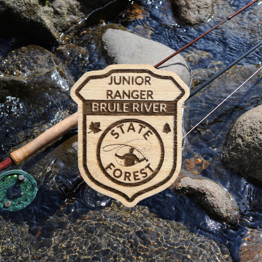 Wood Jr Ranger badge with the text: “Junior Ranger Brule River State Forest” and an icon of a fly fisherman casting.  Photo background: two fly rods laying on the rocks in a stream.