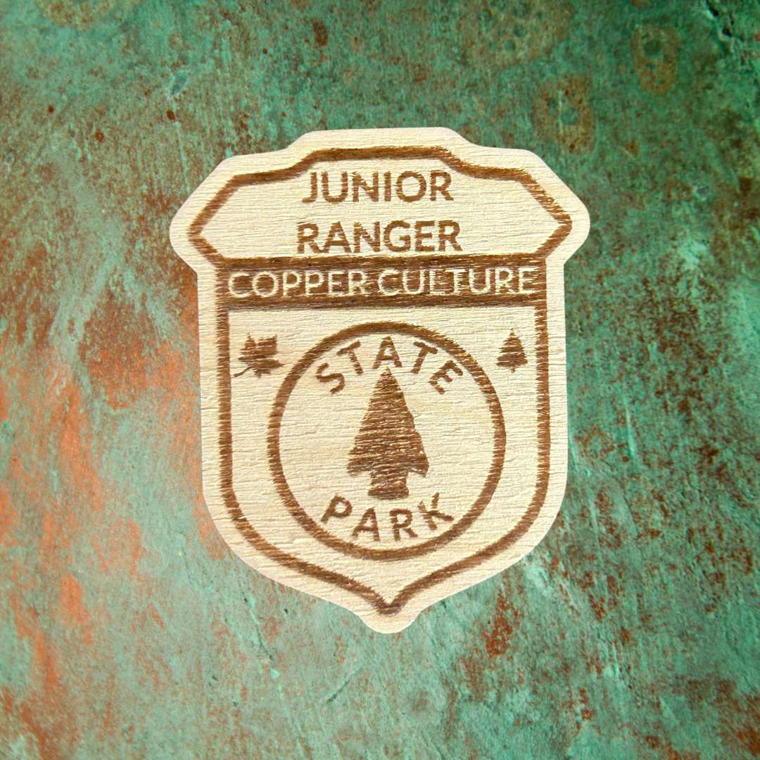 Wood Shaped badge with the text “Junior Ranger Copper Culture State Park” and an arrowhead symbol. Photo background: patina copper metal.