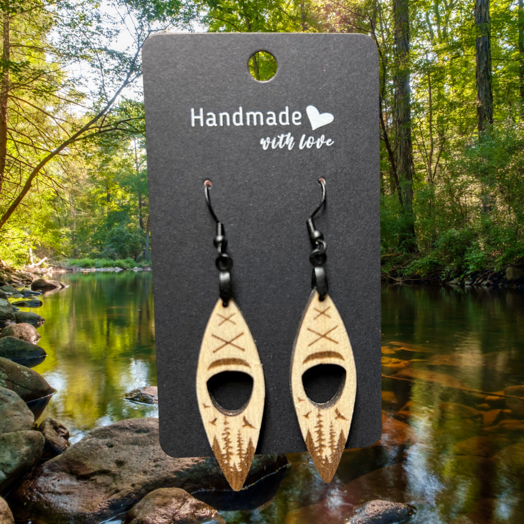 Wood kayak shaped earrings with engraved forest and birds scene. Photo background: stream through a forest.