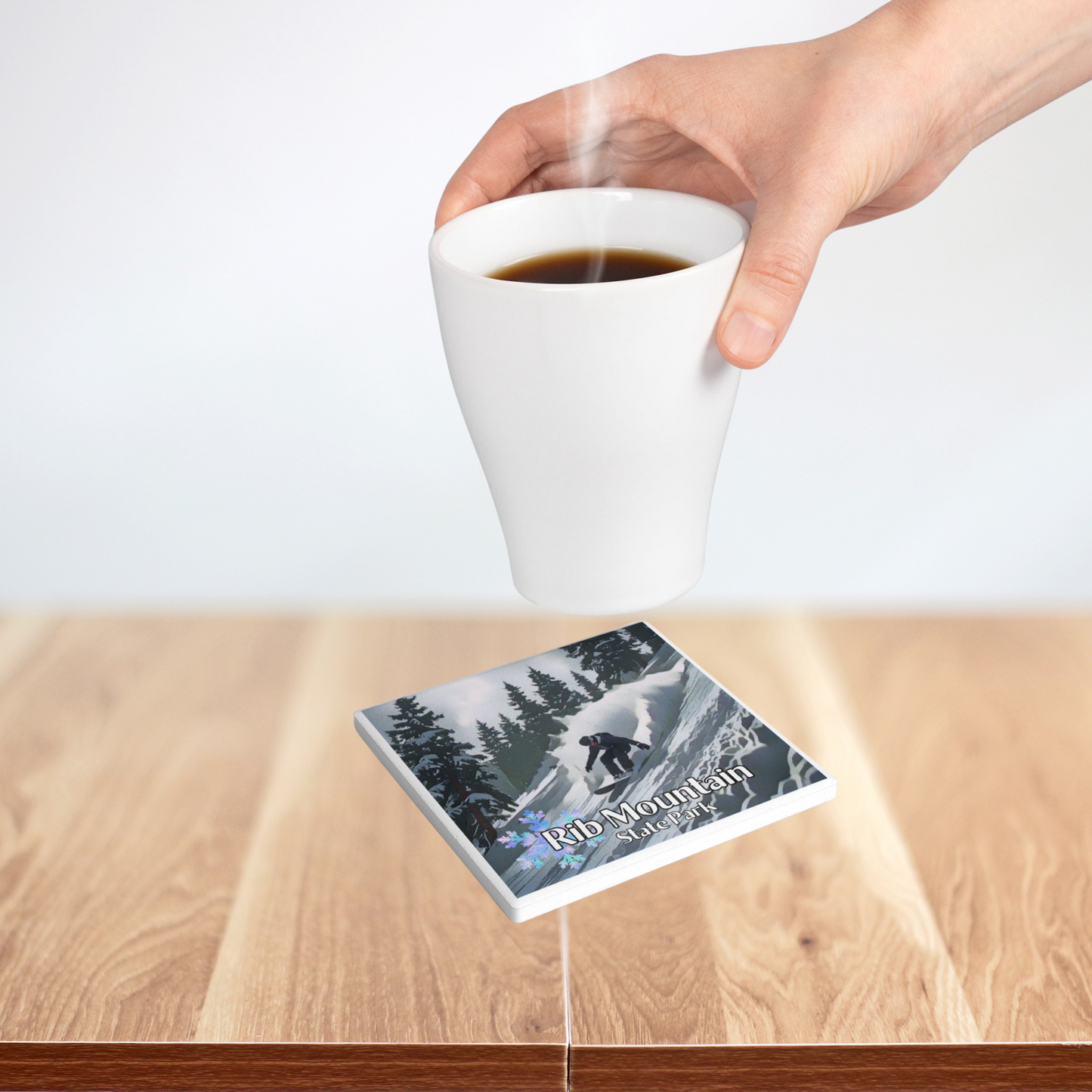 Ceramic coaster featuring a snowboarder going down a snowy, wooded slope and the text: Rib Mountain State Park. Photo Background: person setting a hot beverage on the coaster.