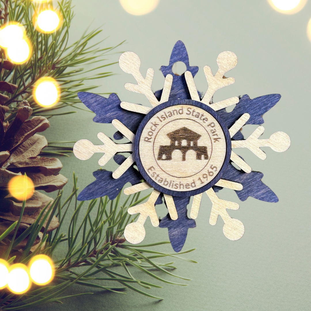 Blue snowflake wood ornament depicting the boat house at Rock Island State Park. Photo background: evergreen branch with lights and a pinecone.