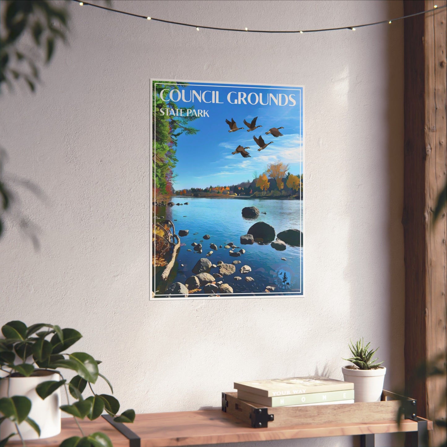 Poster depicting a flock of geese flying over a river lined with fall colored foliage. Photo background: poster on a wall above a shelf with books and potted plants.