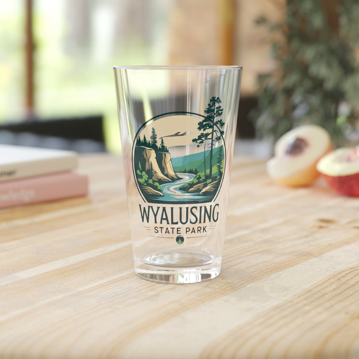 Glass pint glass featuring a design of a winding river below a bluff. Photo Background: glass sitting on a wood table with books and fruit.