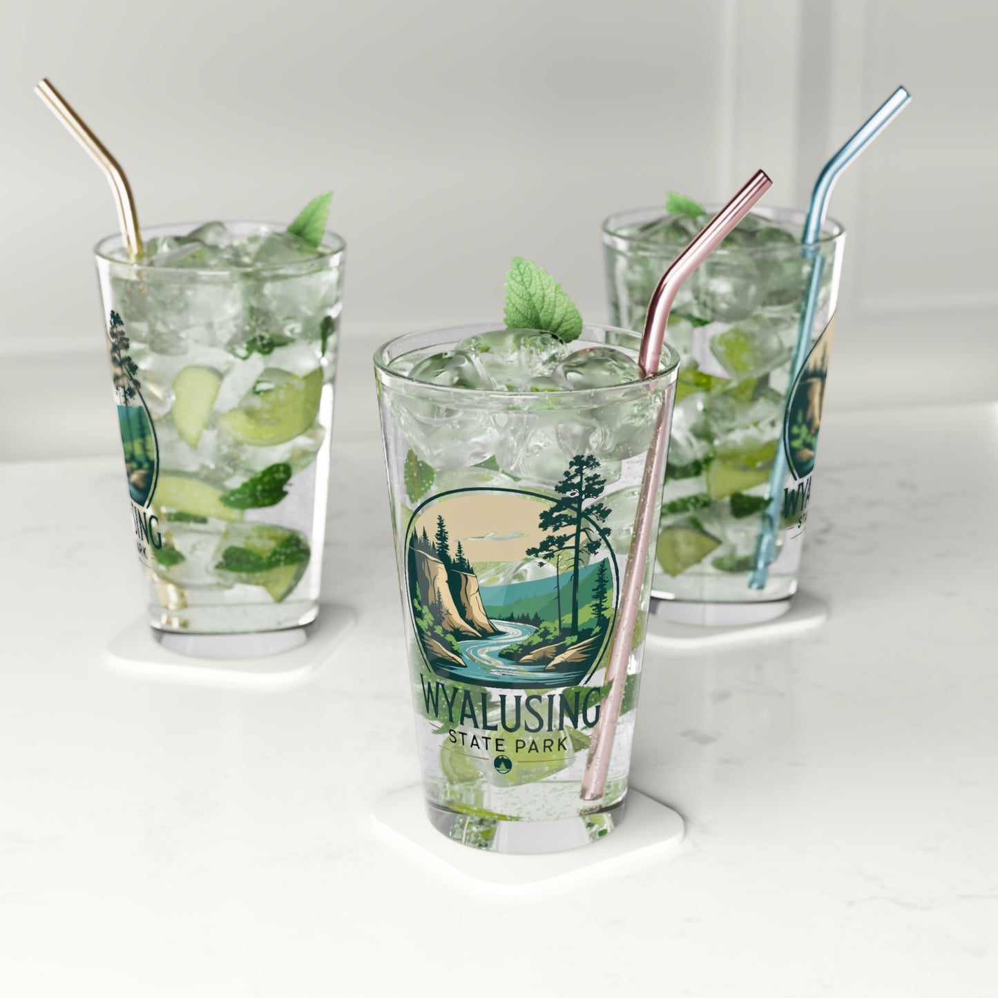 Glass pint glass featuring a design of a winding river below a bluff. Photo Background: three glasses sitting on a counter filled with water and metal straws.