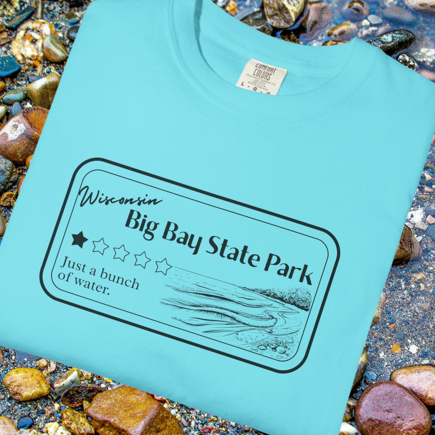 A folded Lagoon Blue Colored T-Shirt with 1 star review design of Big Bay State Park. Features a line art image of a sandy beach and shoreline, 1 star graphic, and the text: “Wisconsin Big Bay State Park, Just a bunch of water. T-shirt is folded and sitting on rounded lake pebbles.