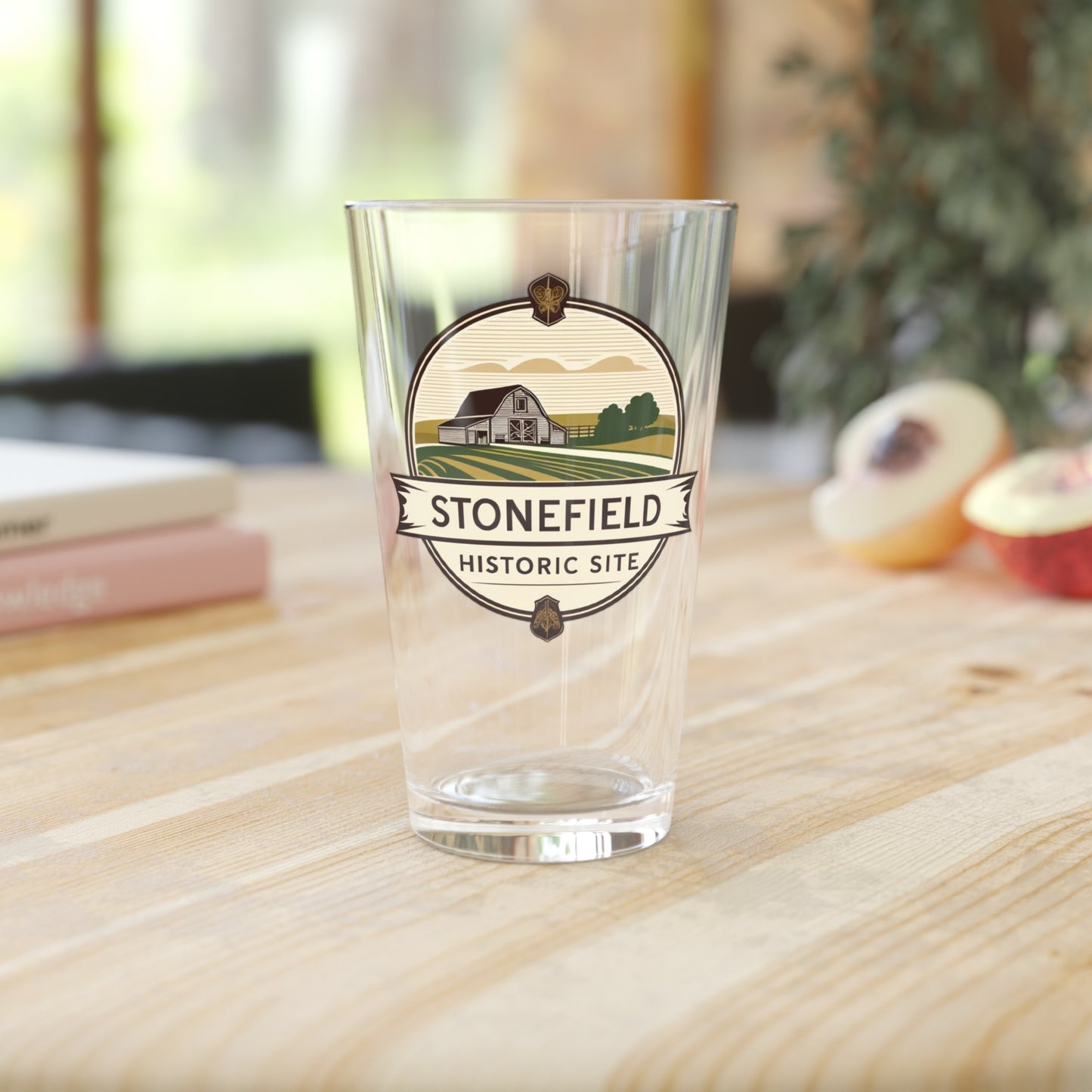 16oz pint glass featuring a logo design of a barn set amongst fields. Photo background: glass sitting in a wood counter with books and fruit.