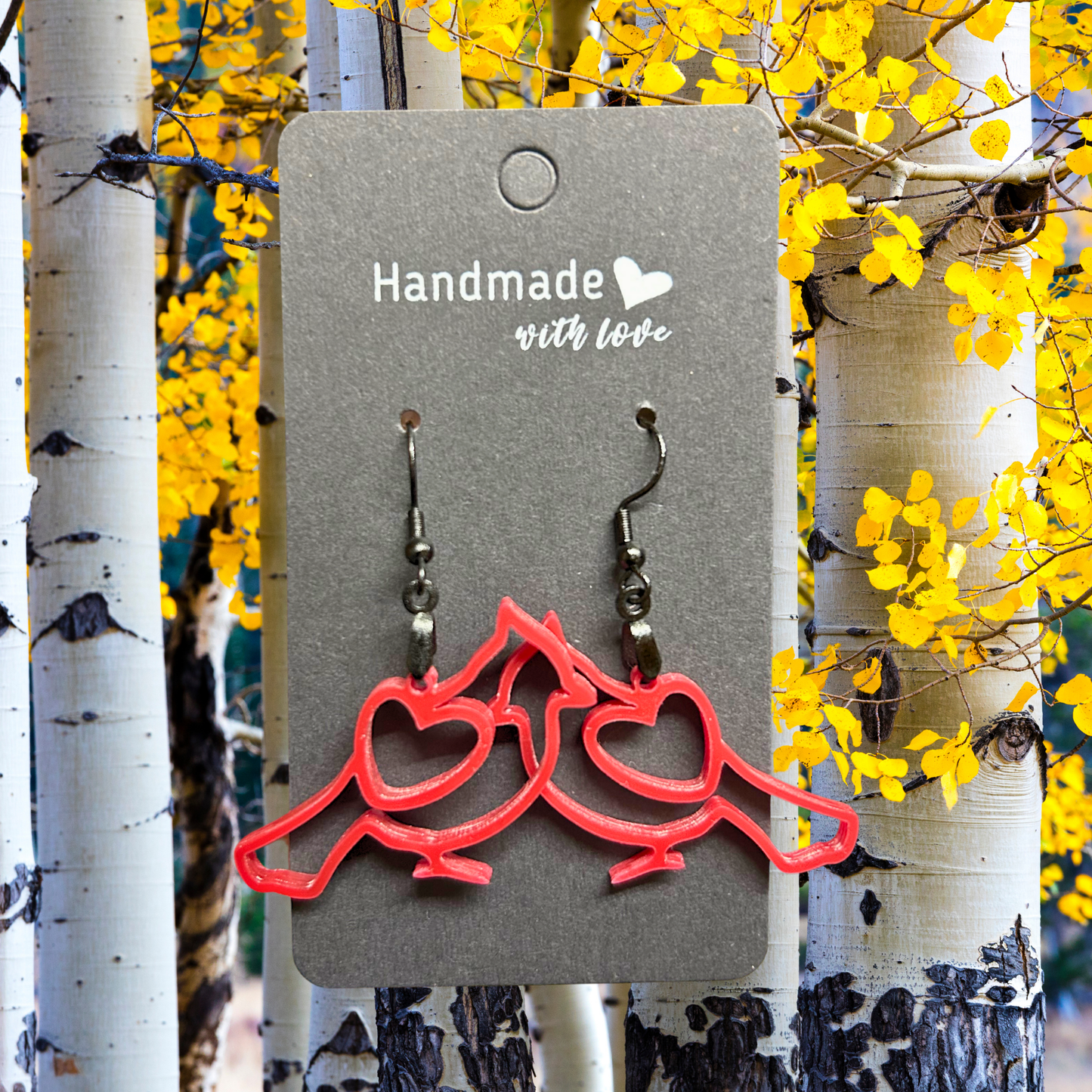Pair of red cardinal earrings on a black hangtag. Background of aspen trees and yellow leaves.