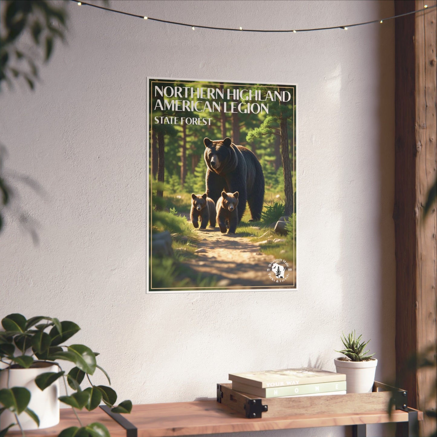 Poster of a Black bear and two cubs walking through a pine forest and the text: Northern Highland American Legion State Forest. Photo Background: poster hanging on a wall above a wood shelf with books and potted plants.