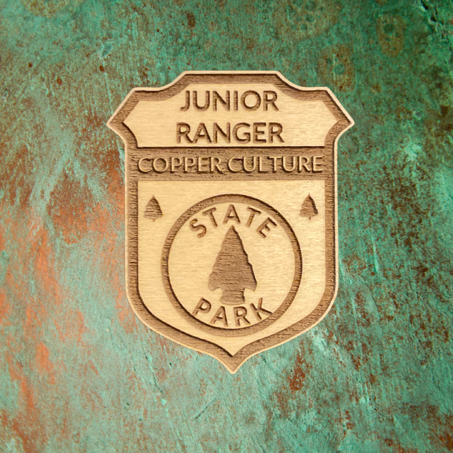Wood Shaped badge with the text “Junior Ranger Copper Culture State Park” and an arrowhead symbol. Photo background: patina copper metal.