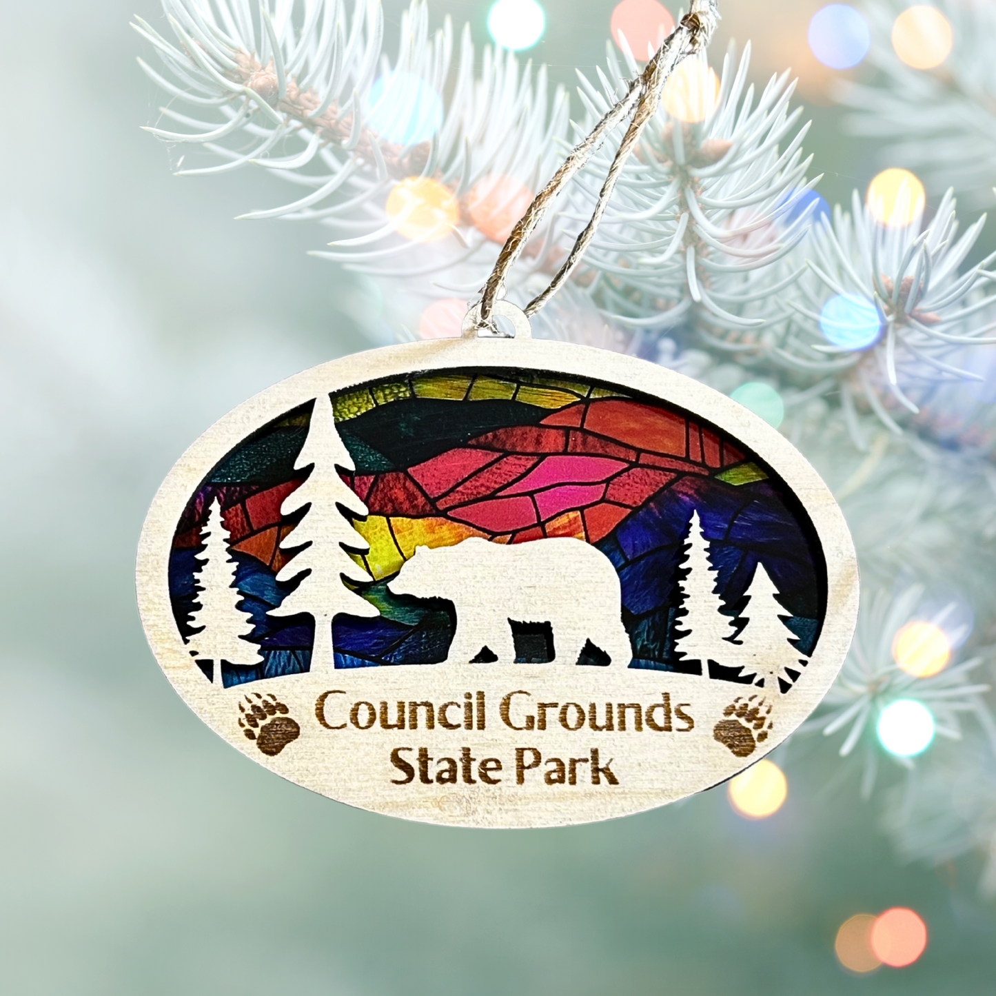 Oval wood bear ornament featuring a bear and trees and the text Council Grounds State Park with a faux stained glass background. Photo background: close up photo of a tree branch with Christmas lights.