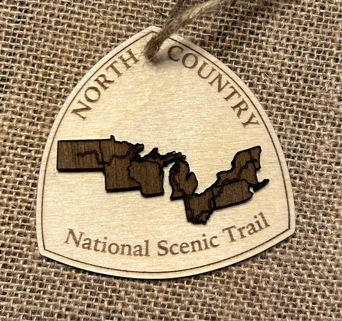 Wood ornament in the shape of the national trail's icon, with a walnut-colored North Dakota, Minnesota, Wisconsin, Michigan, Ohio, Pennsylvania, New York, and Vermont Shaped center piece depicting the North Country Trail Route. Ornament contains the text "North Country National Scenic Trail".