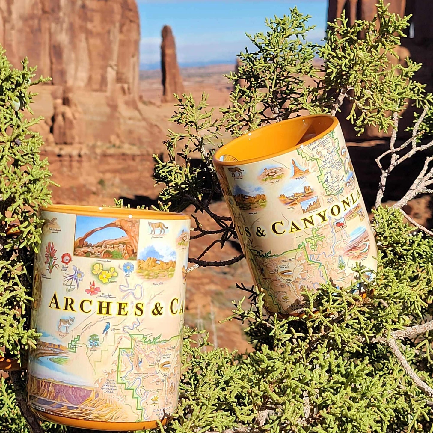Two cylindrical mugs printed with a colorful illustrated map of Arches National Park, placed among green desert shrubs with a canyon rock formation in the background.