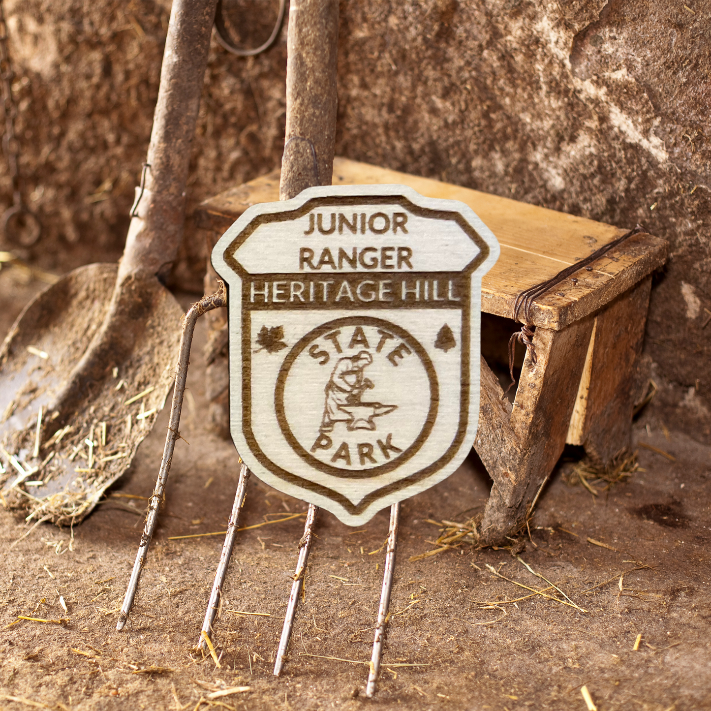 Wood badge shaped JR Ranger Badge with the text “Junior Ranger, Heritage Hill State Park” with a blacksmith symbol. Photo background: old shovel and pitchfork leaning against a wall, hay laying on the floor.