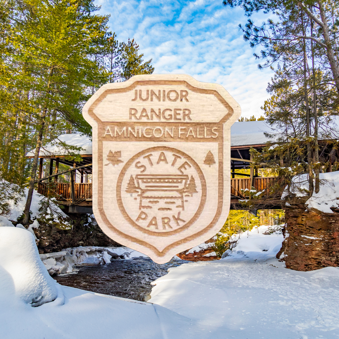 Wood shaped Jr. Ranger Badge depicting a covered bridge over a stream and the text: Junior Ranger Amnicon Falls State Park. Photo Background: covered bridge over a partially frozen river.