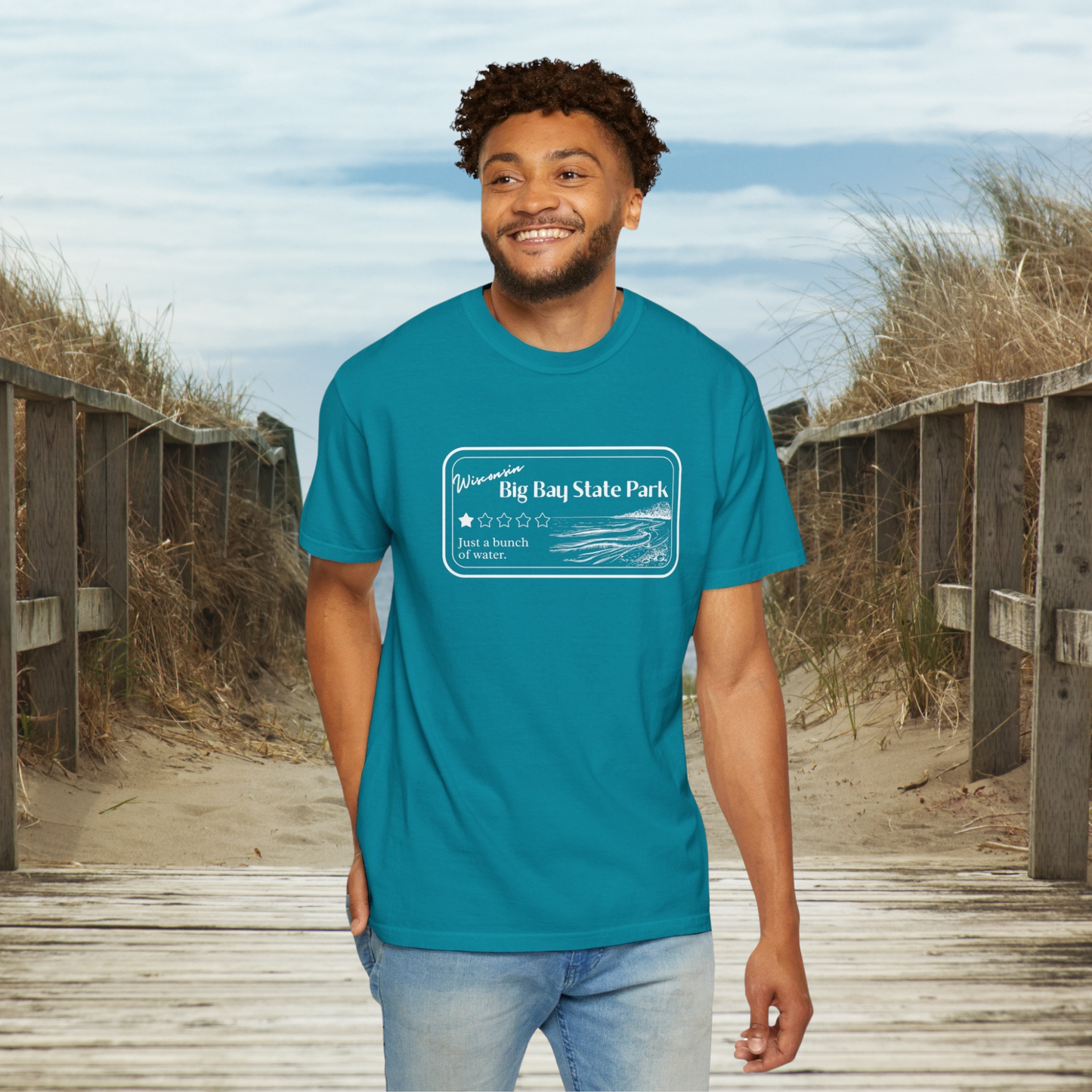 Male model standing a boardwalk to a beach wearing a Topaz Blue Colored T-Shirt with 1 star review design of Big Bay State Park. Features a line art image of a sandy beach and shoreline, 1 star graphic, and the text: “Wisconsin Big Bay State Park, Just a bunch of water.