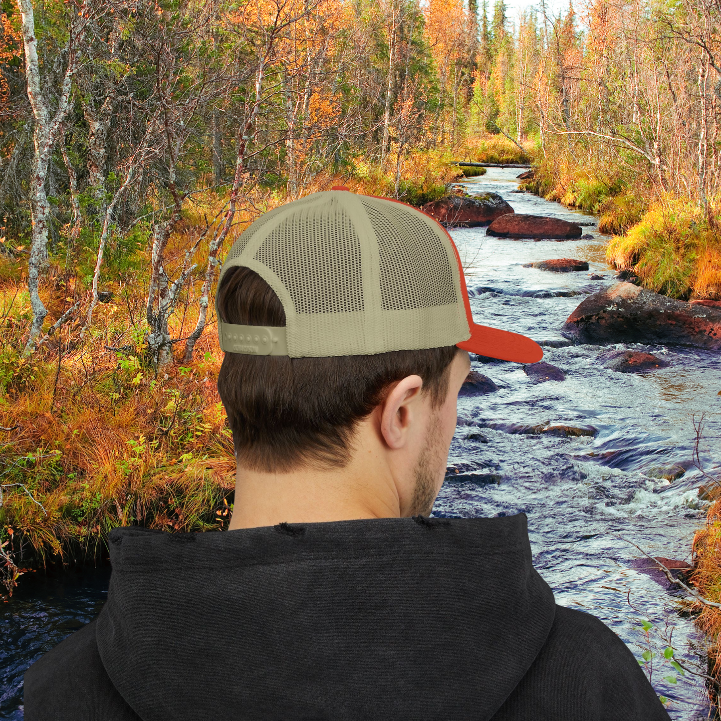 Male model wearing a Rustic Orange and Khaki Colored ball cap with embroidered logo of an angler catching a fish and the text Brule River State Forest. Model is looking away at stream running through woods in fall.