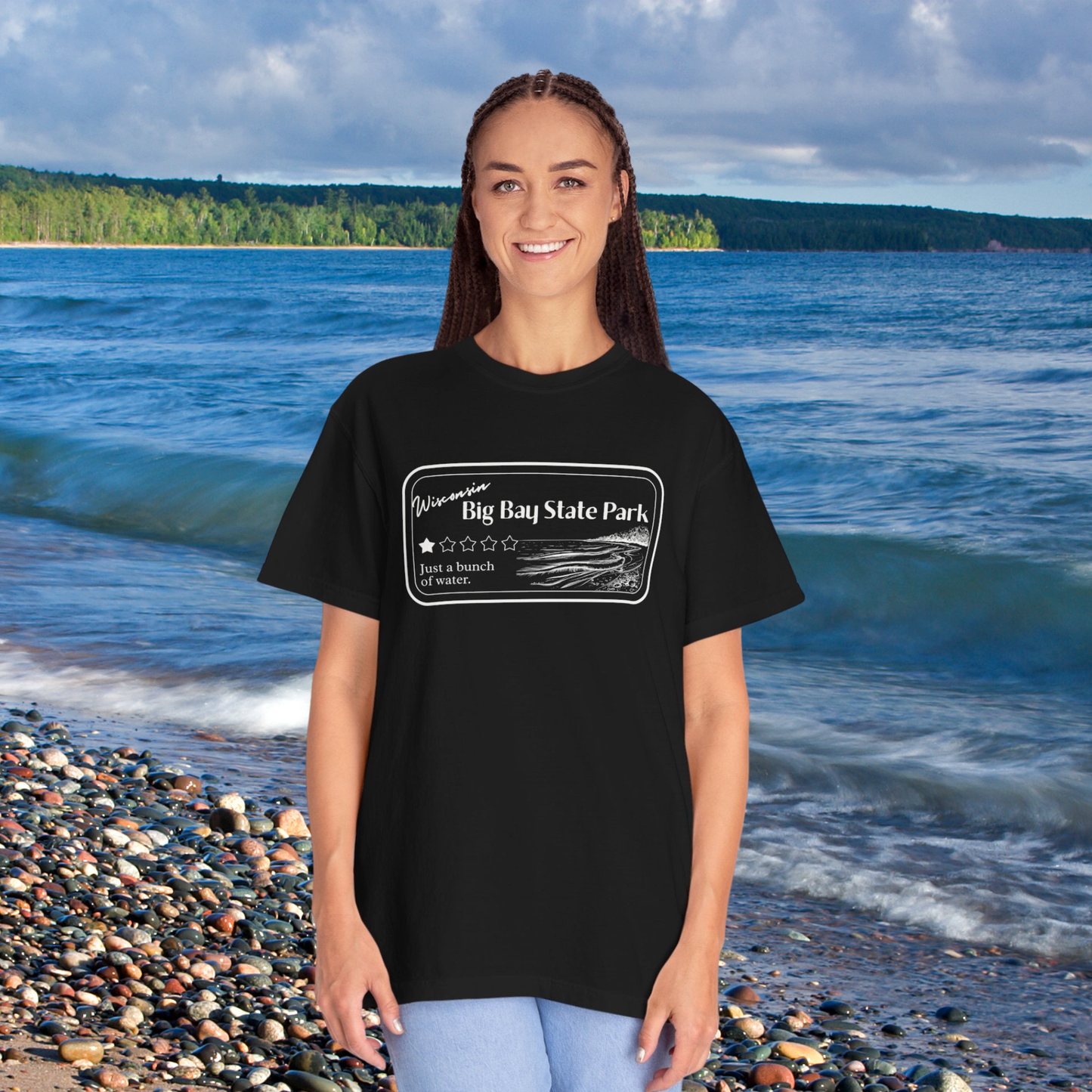 Female standing on a rock beach with water in the background wearing a Black Colored T-Shirt with 1 star review design of Big Bay State Park. Features a line art image of a sandy beach and shoreline, 1 star graphic, and the text: “Wisconsin Big Bay State Park, Just a bunch of water.
