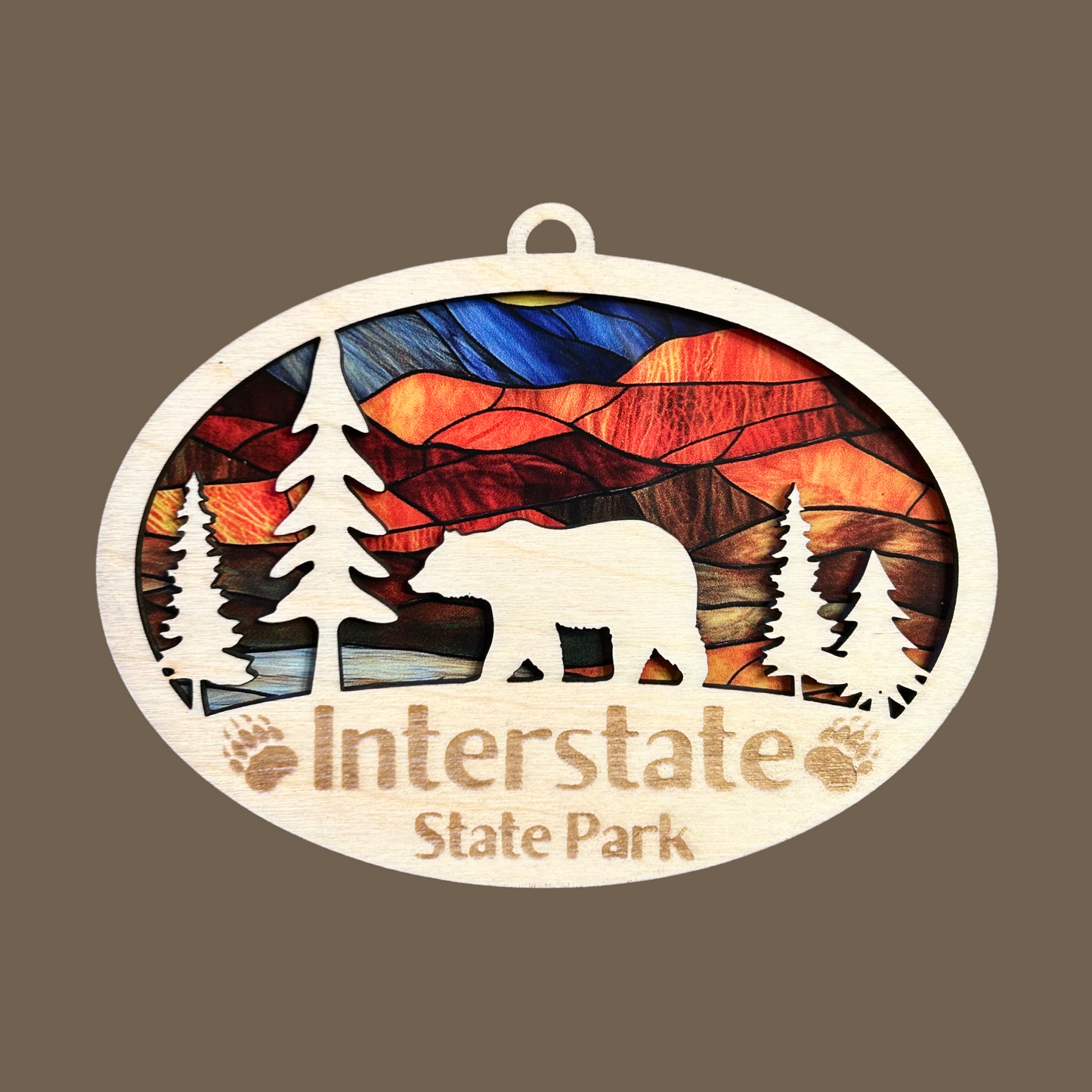 Oval shaped ornament with trees and bear and a stained glass appearance. Includes the text “Interstate State Park” with engraved paw prints. Photo Background: brown.