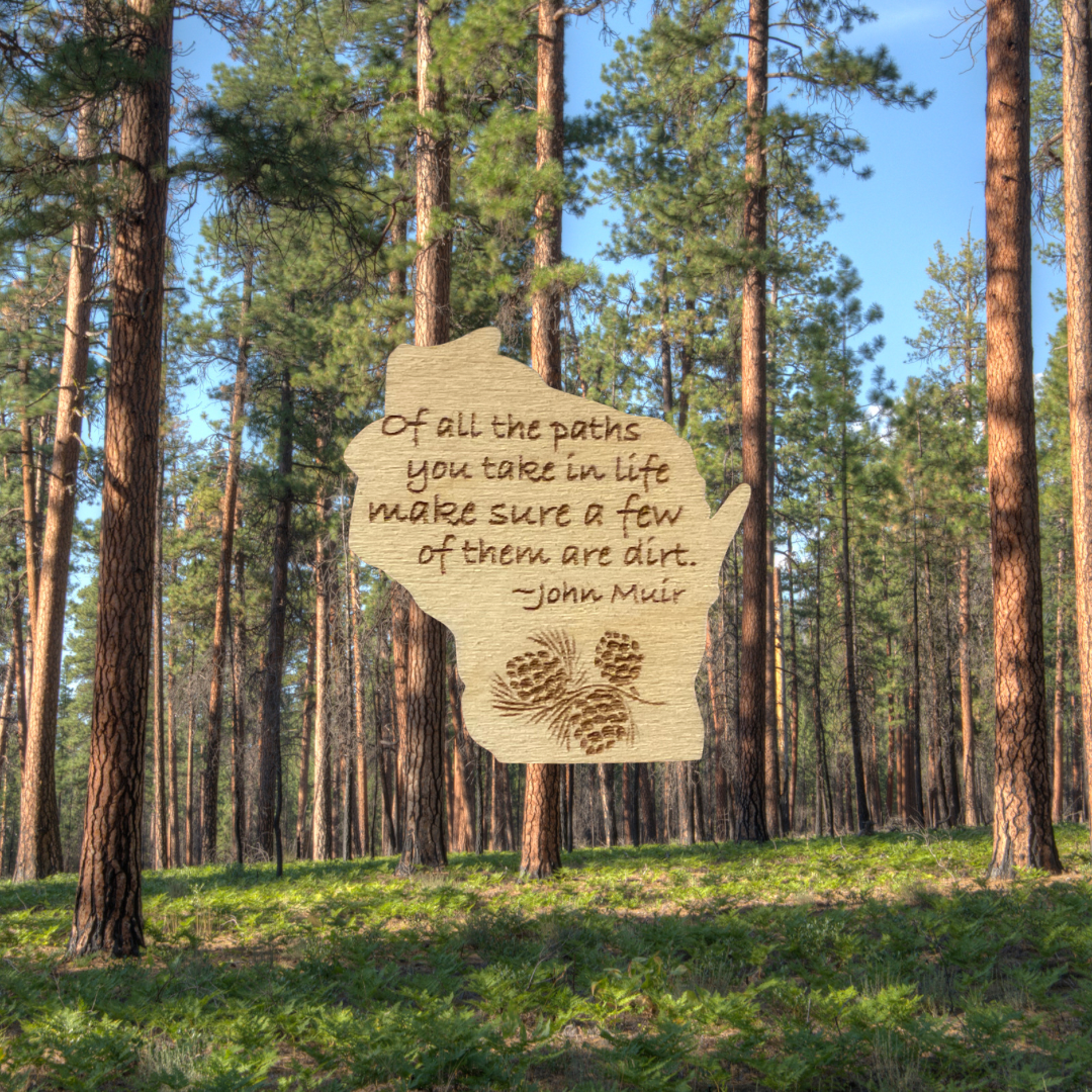 Wood Wisconsin shaped magnet with the John Muir quote: “of all the paths you take in life, make sure a few of them are dirt.” Background of tall pine trees.