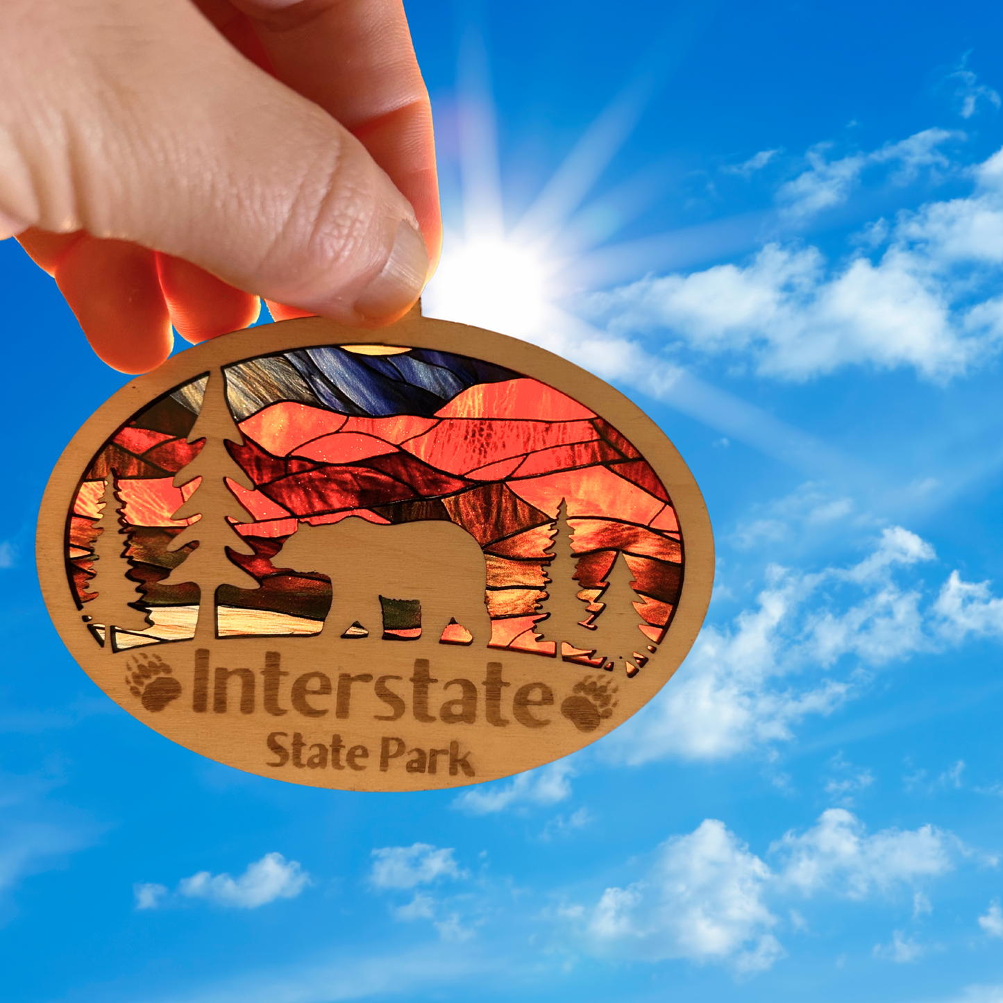 Oval shaped ornament with trees and bear and a stained glass appearance. Includes the text “Interstate State Park” with engraved paw prints. Photo Background: ornament held up to the sun to show it’s vibrant colors.
