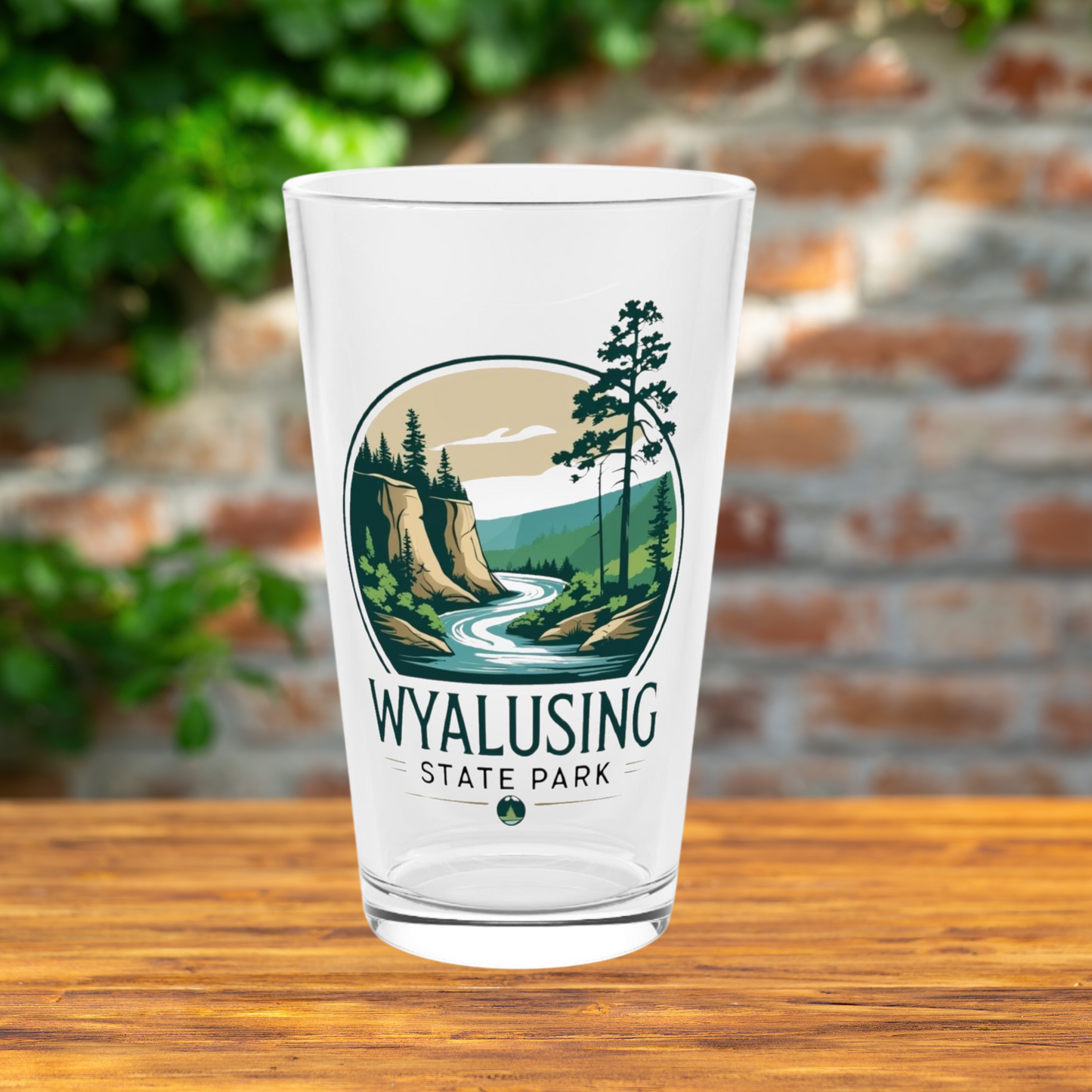Glass pint glass featuring a design of a winding river below a bluff. Photo Background: glass sitting on a wood table in front of a brick wall.