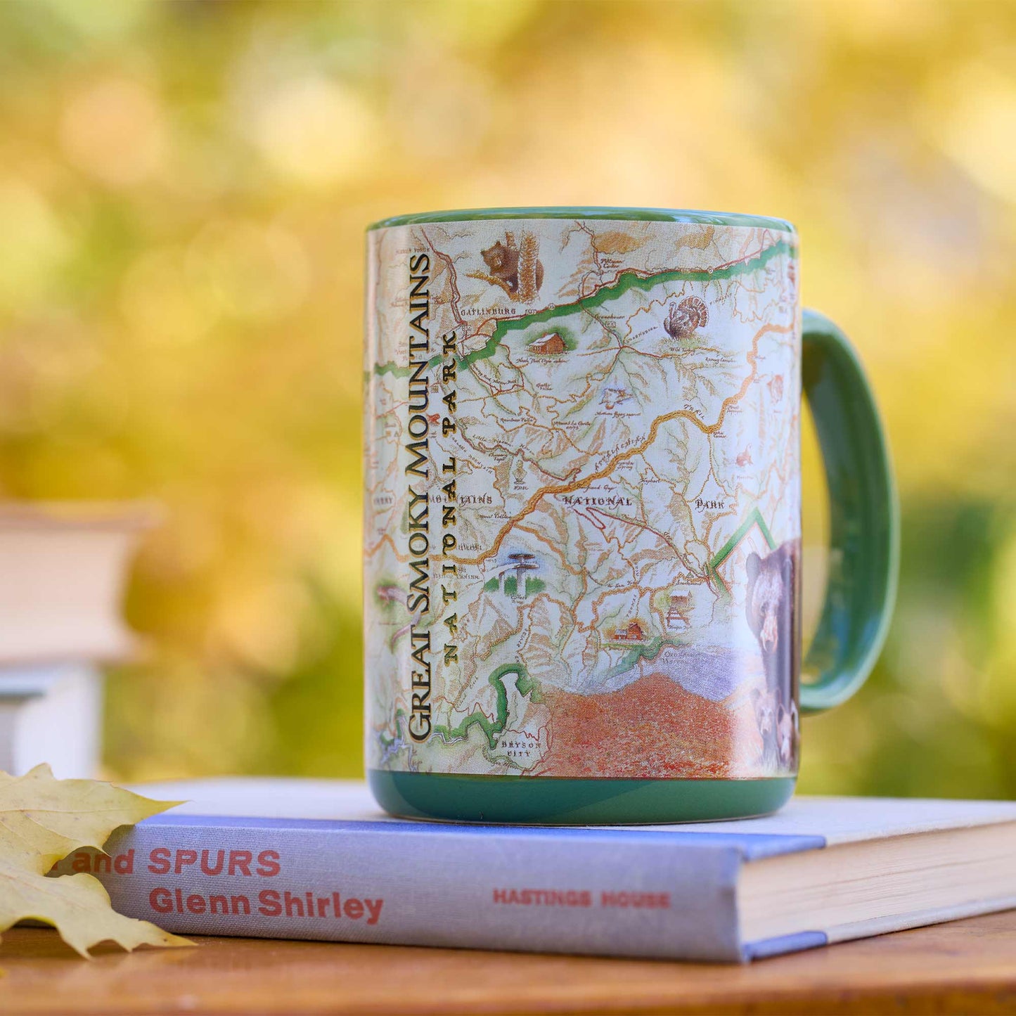 A ceramic mug printed with a vintage-style map of Great Smoky Mountains National Park, resting on a book with a leaf in front, against a blurred autumn outdoor background.