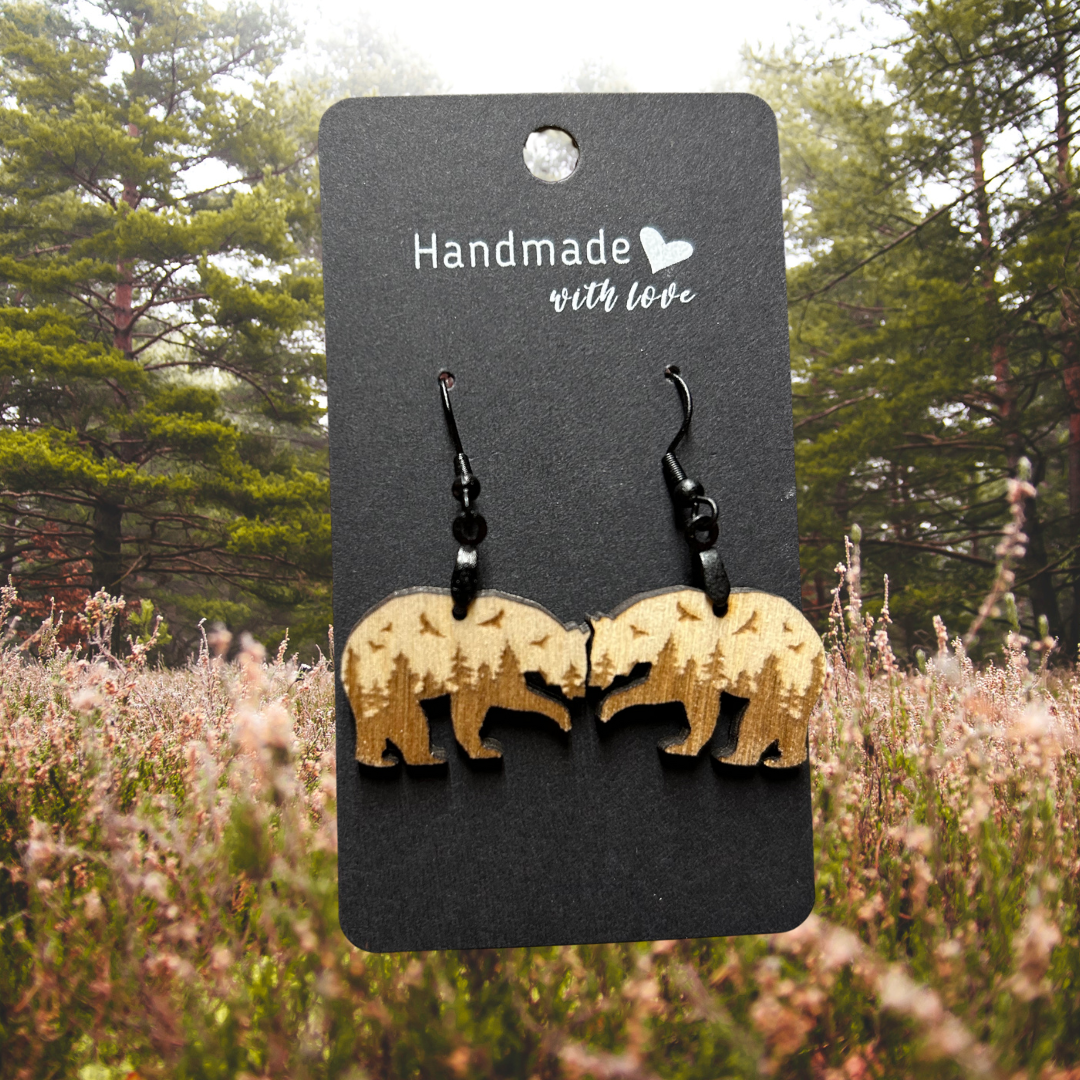 Bear shaped wood earrings with forest scene and flying birds engraved on them. Photo background: pine trees lining a misty meadow.