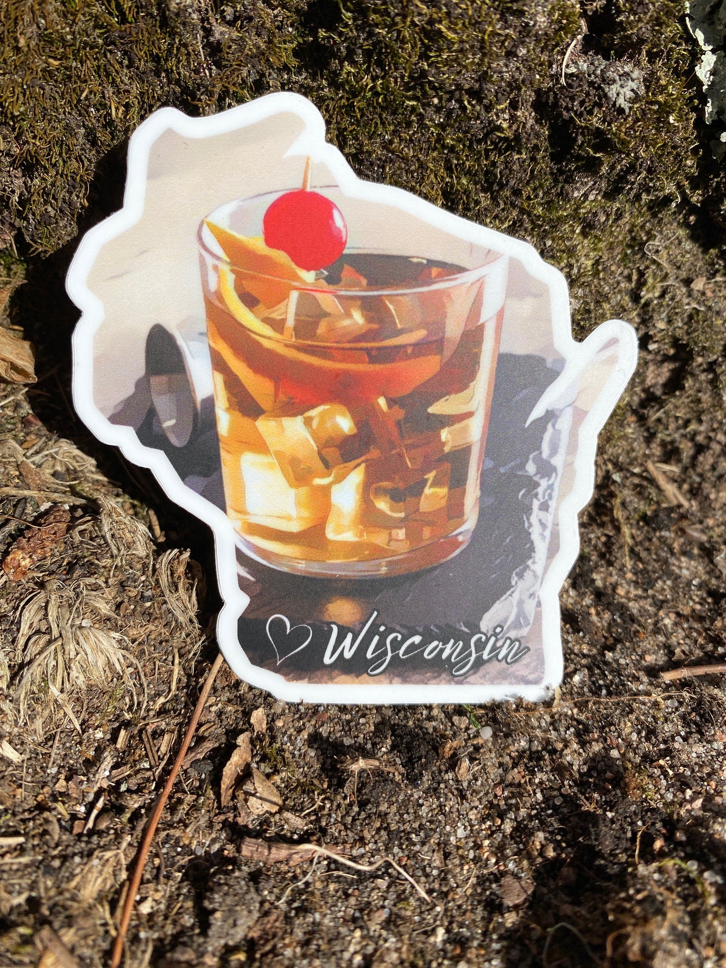 Wisconsin shaped sticker featuring a rendering of an old fashioned drink on a coaster. Photo Background: dirt and grass.