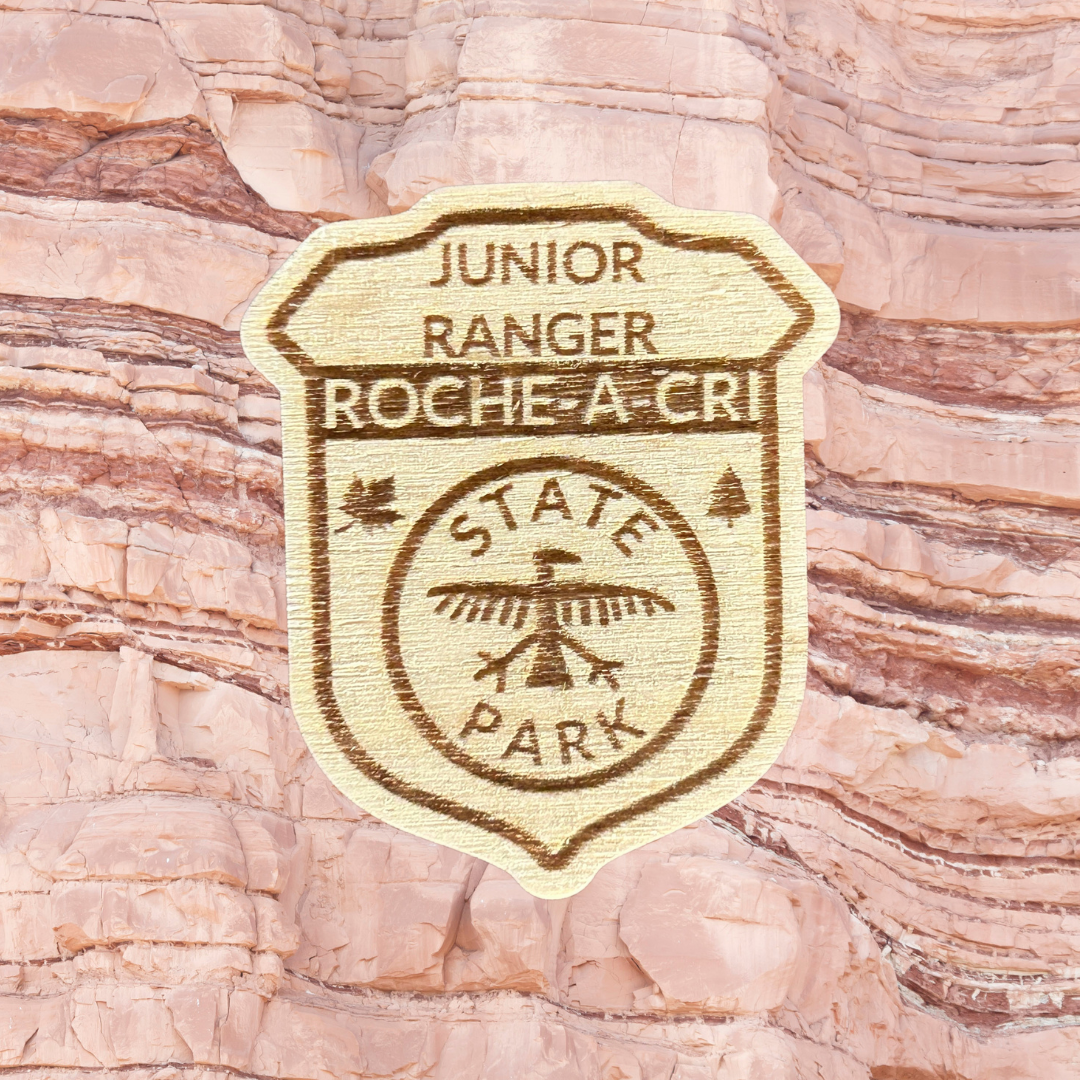 Wood Shaped badge with the text “Junior Ranger Roche-A-Cri State Park” and an petroglyph symbol. Photo background: rock.